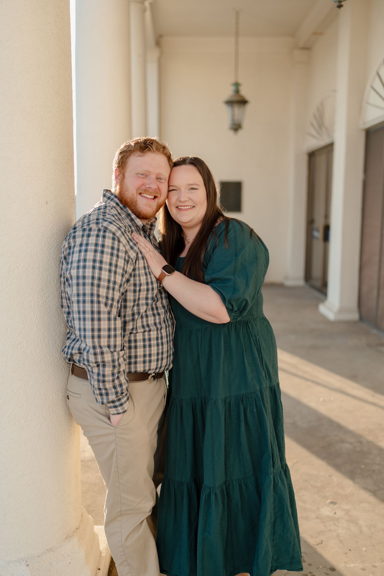 Couple posing against white column, woman in green dress embracing man in plaid shirt, warm golden hour lighting.