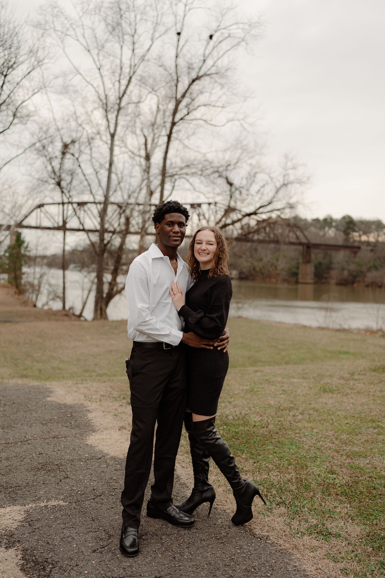 Smiling interracial couple embracing outdoors near river and iron bridge, woman in black dress and tall boots.