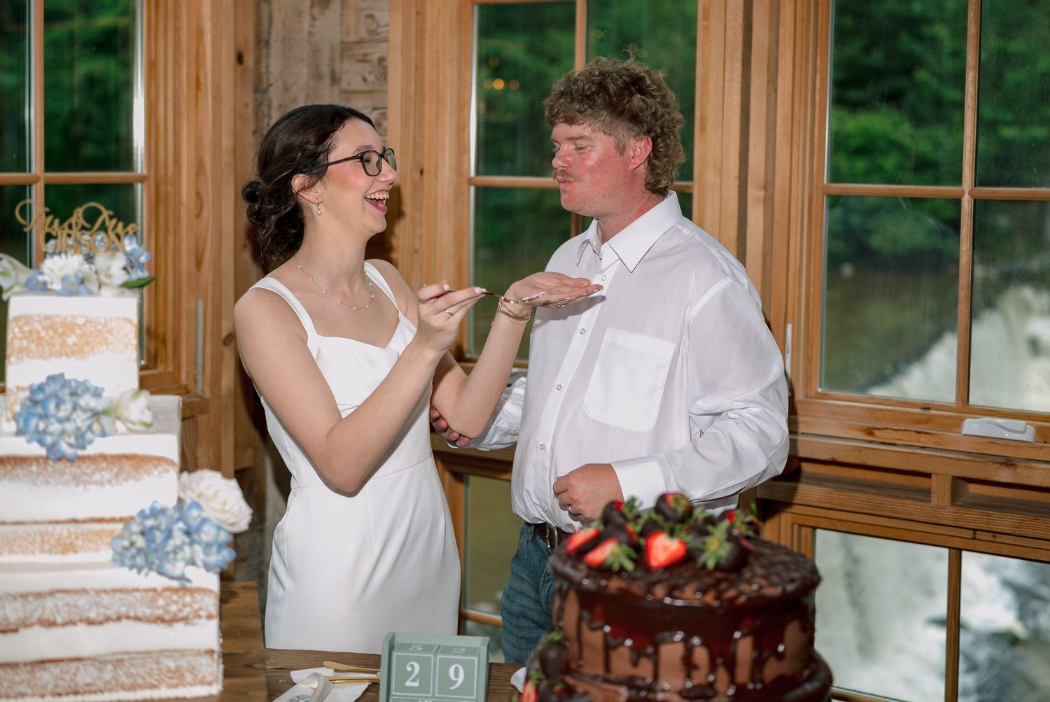 Bride playfully feeds cake to groom during wedding reception with elegant tiered cakes in background.