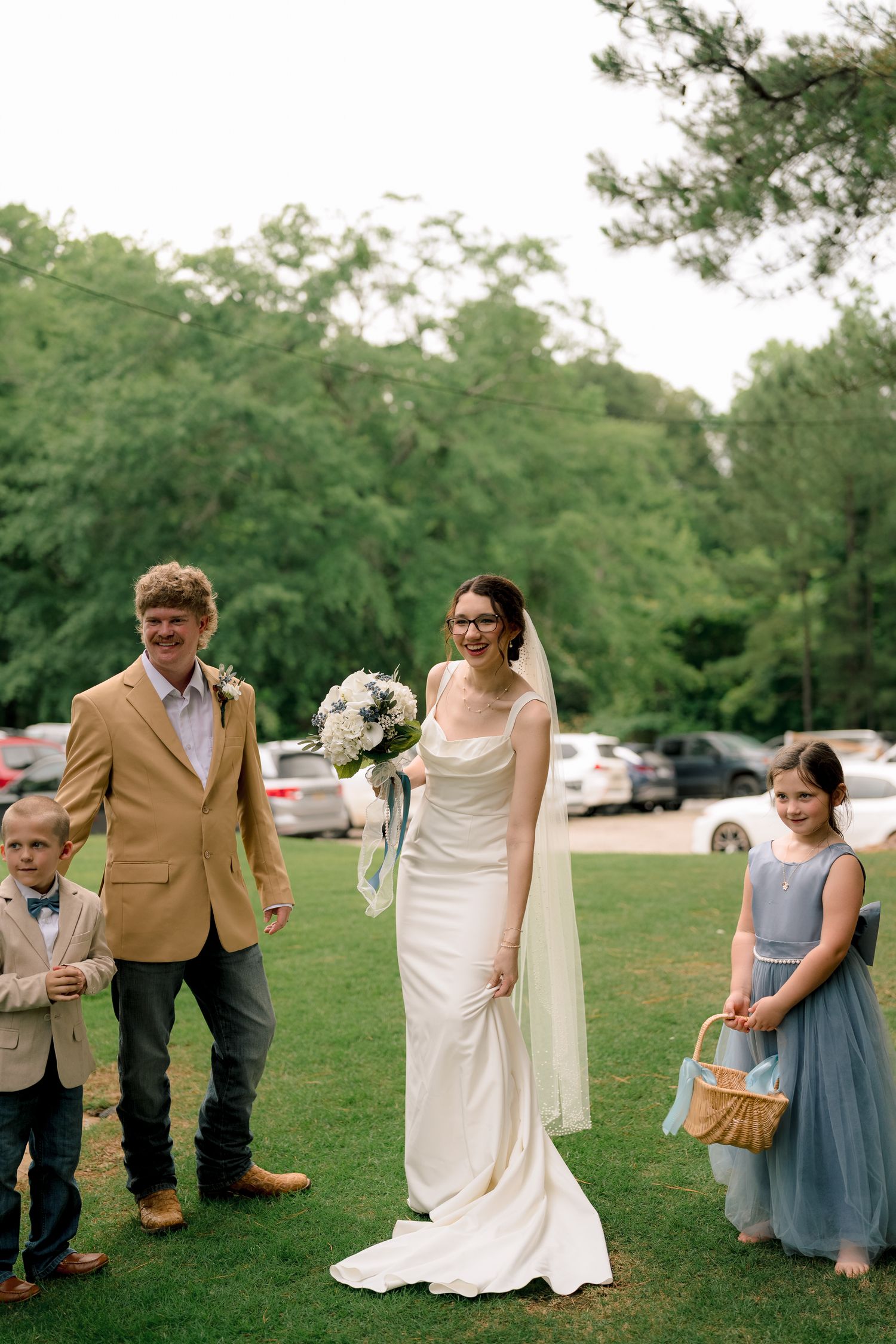 Bride in white gown with groom, ring bearer, and flower girl posing on green lawn.