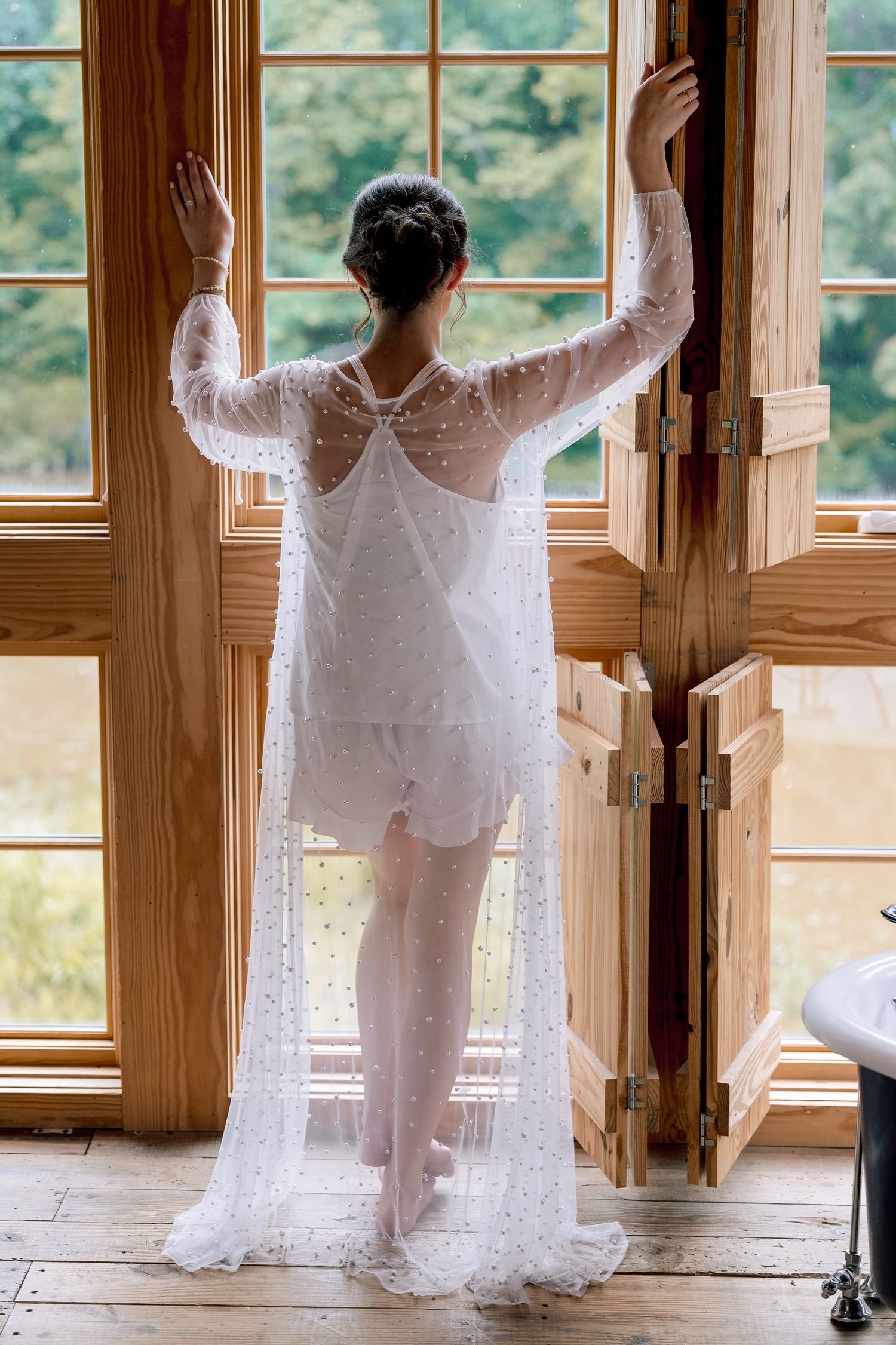 Bride in white pearl-embellished robe with train stands at wooden French doors overlooking lush greenery.