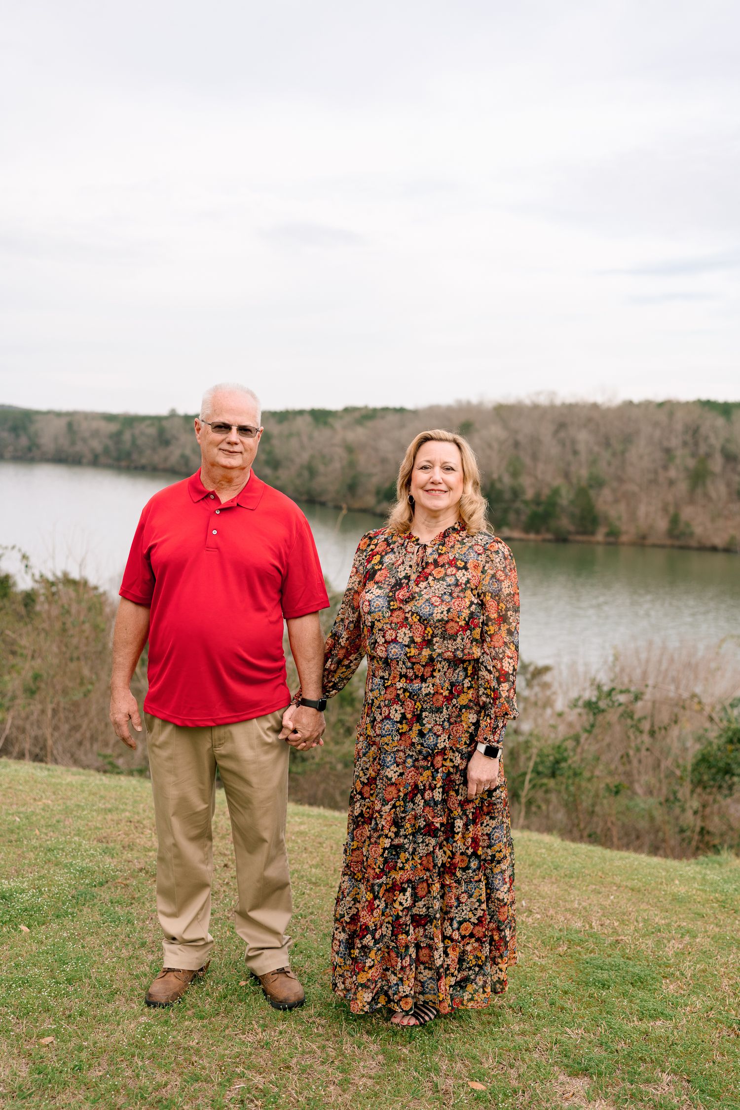 Couple holding hands outdoors by a lake, man in red polo and woman in floral dress, smiling.