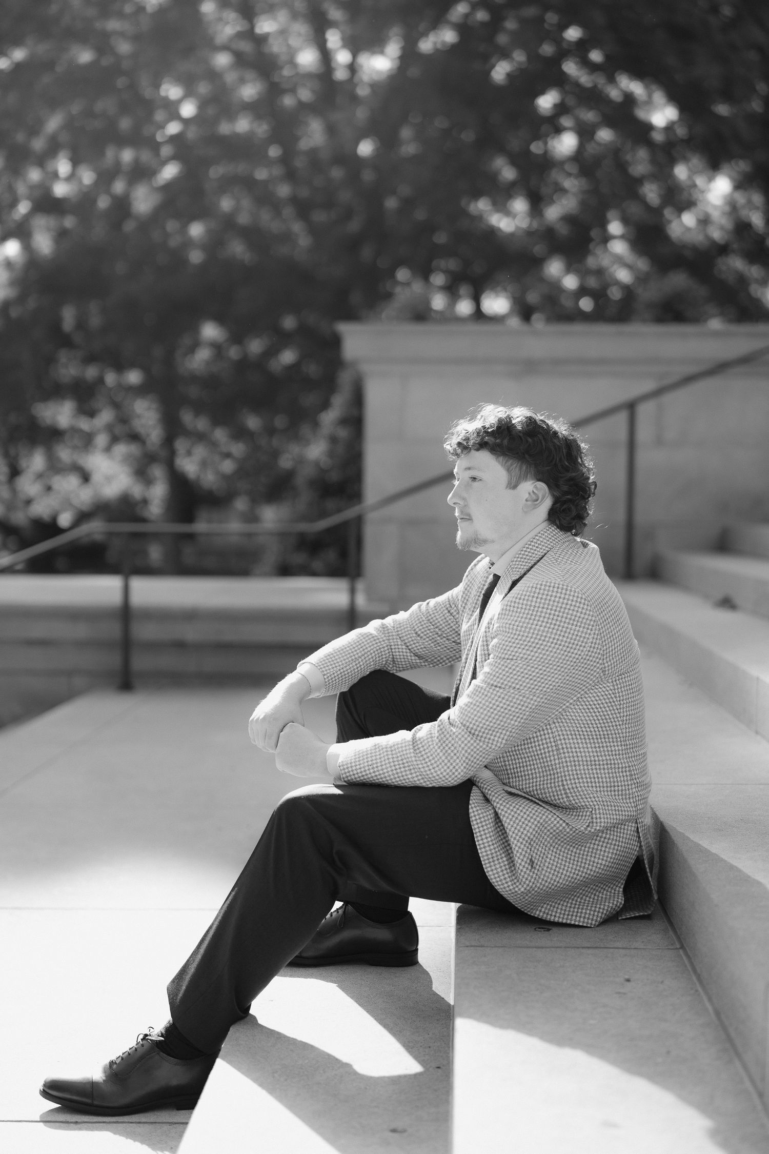 Black and white photo of young man in blazer sitting on stone steps gazing thoughtfully to the side.