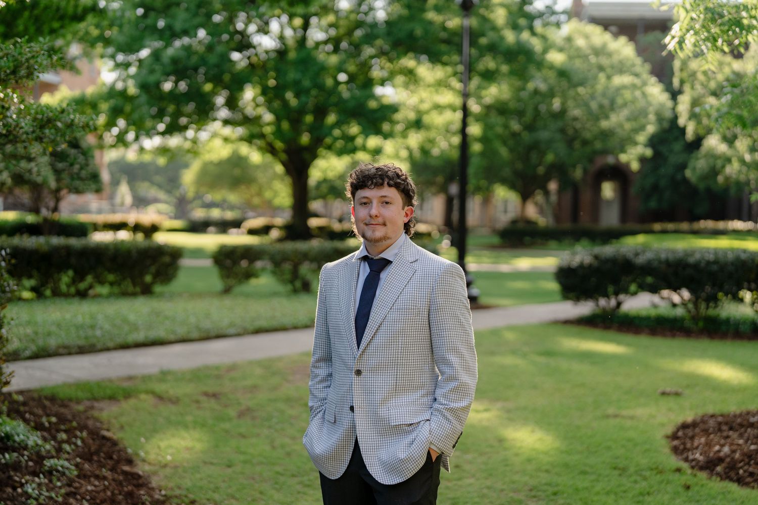 Young man in light blazer and dark pants standing on a tree-lined campus walkway.