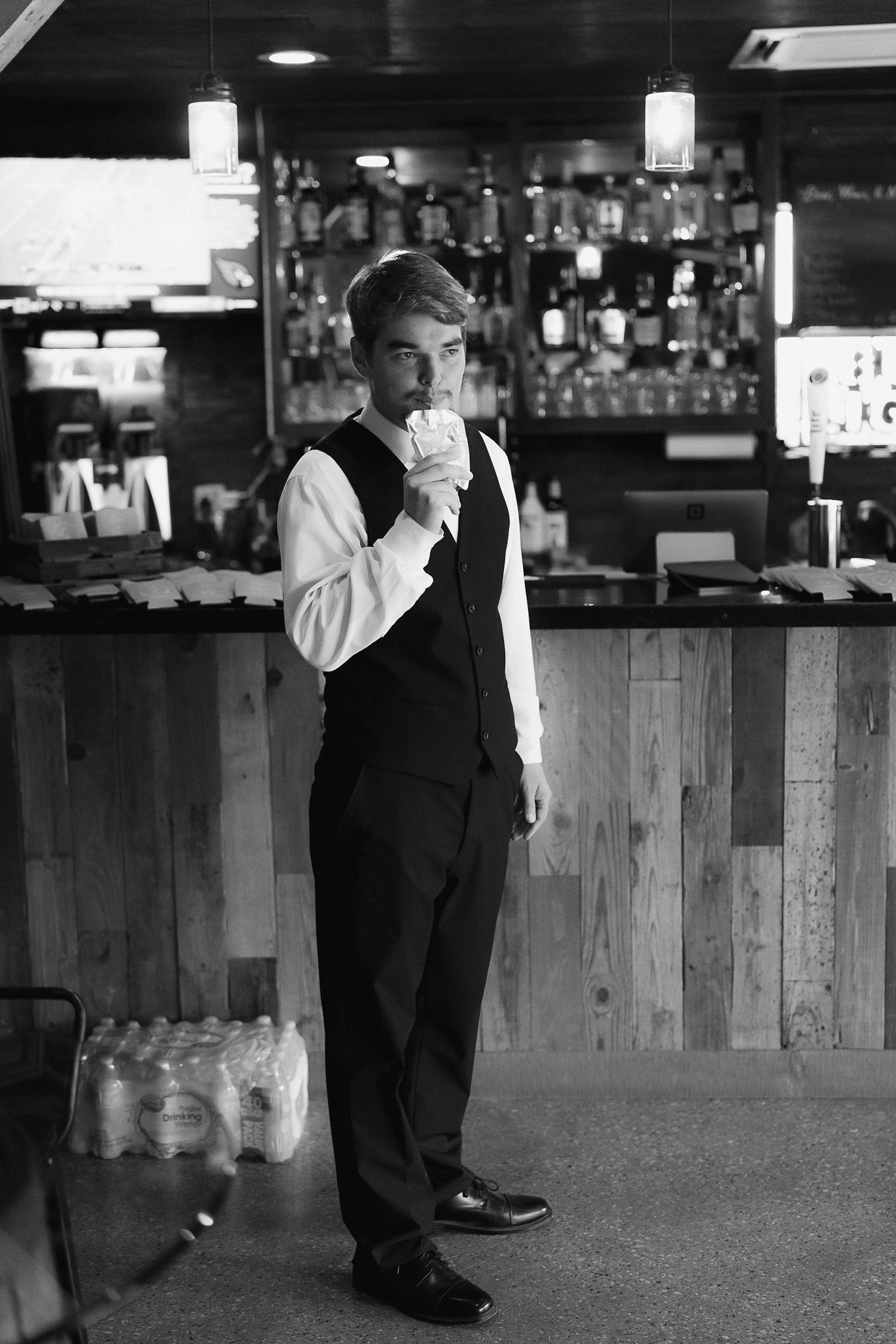 Dapper groom in black vest and white shirt sipping a drink at a rustic bar before his wedding ceremony.