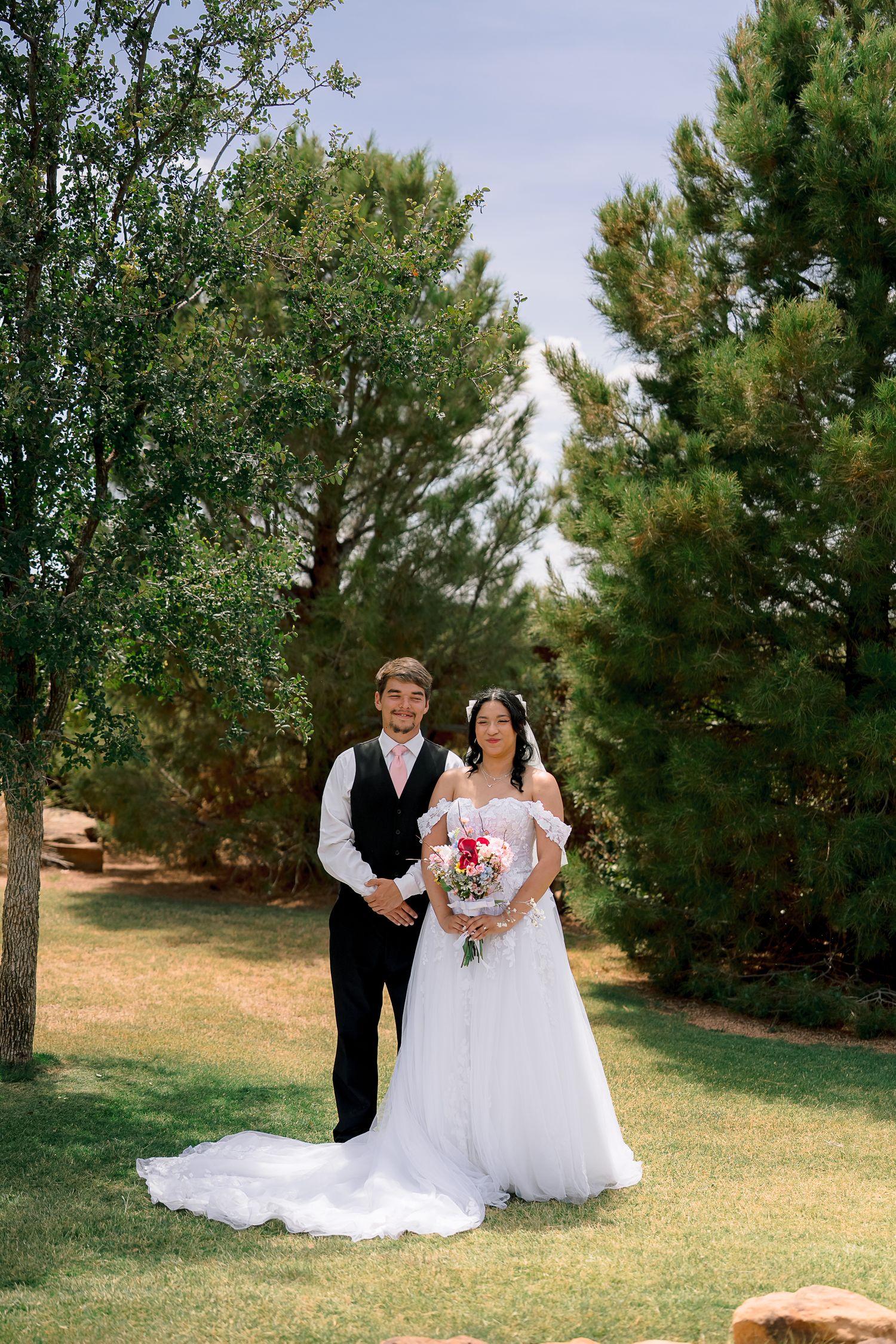 Bride and groom posing together outdoors on green grass surrounded by tall pine trees on their wedding day.
