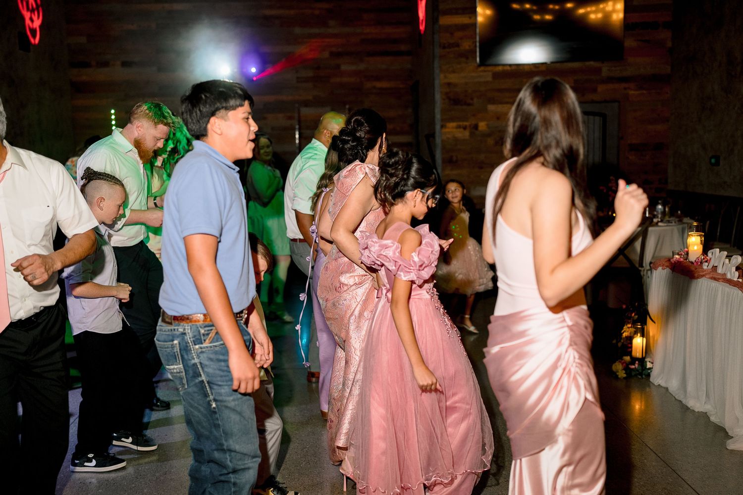 Guests dancing at wedding reception in dimly lit rustic venue with colorful lighting.