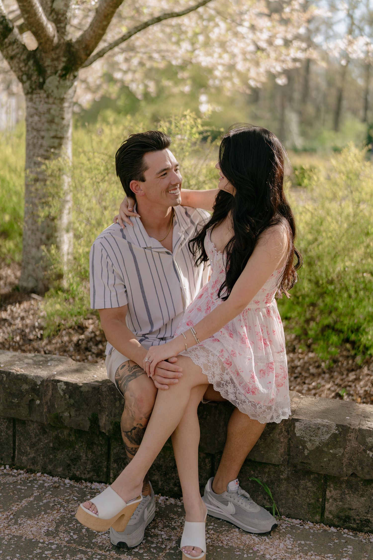 Smiling couple sitting together on a stone wall under blooming spring trees in soft golden light.