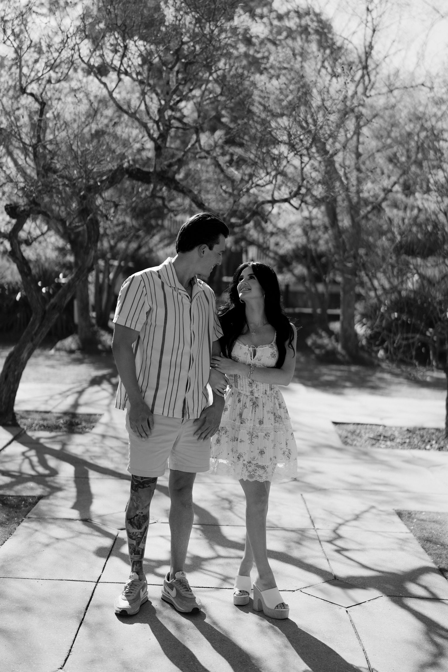 Black and white photo of couple gazing at each other on sunny garden path with tree shadows.
