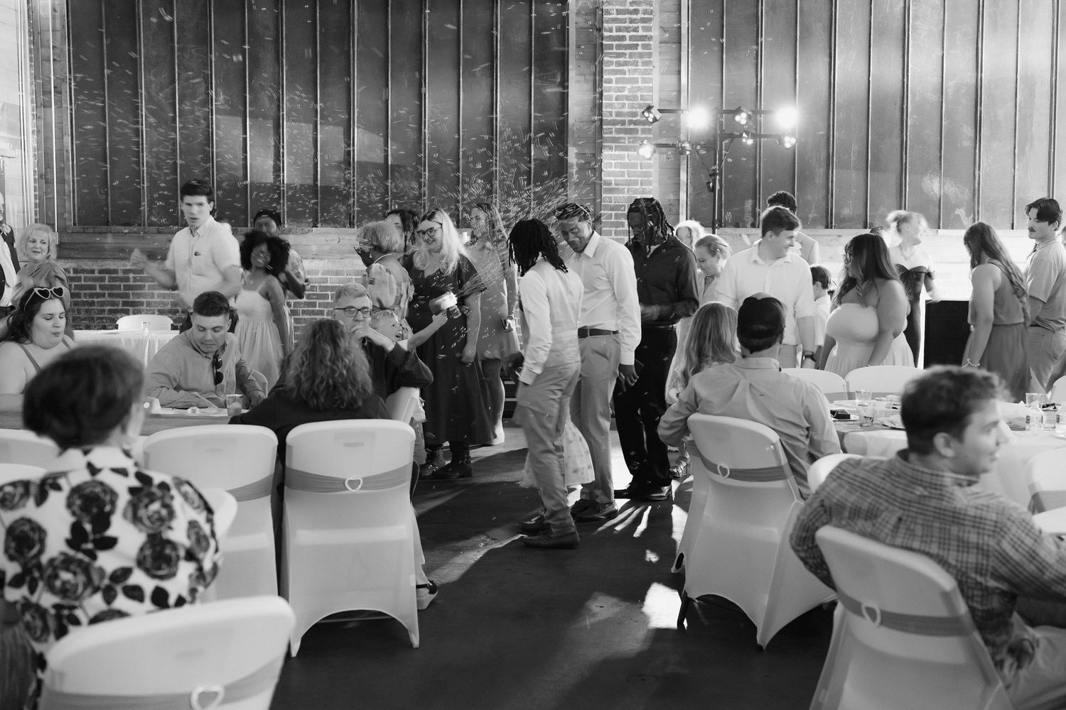 Wedding reception guests mingling and dancing in black and white photo inside a large brick venue hall.