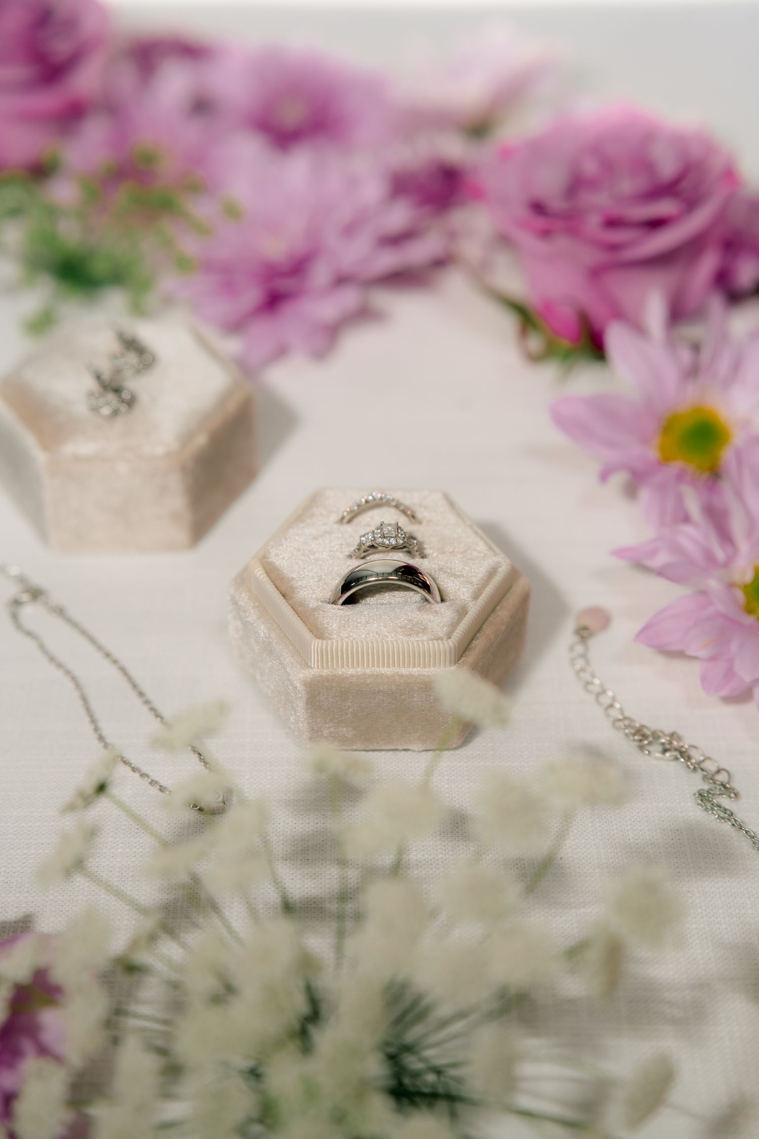 Wedding rings and diamond jewelry displayed in a velvet hexagonal box surrounded by pink and white flowers.