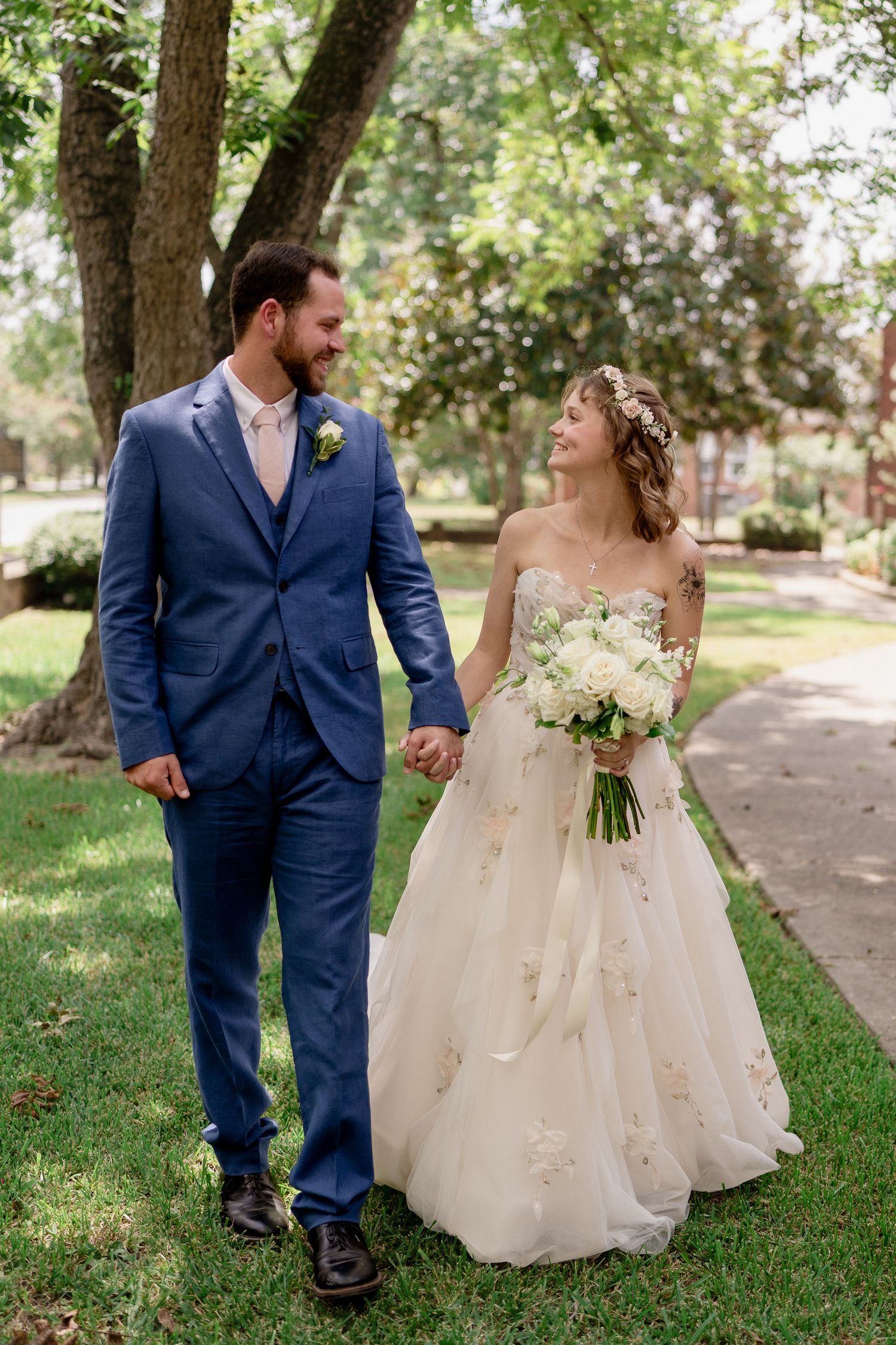 Wedding couple walks hand in hand through a tree-lined path in a garden setting.