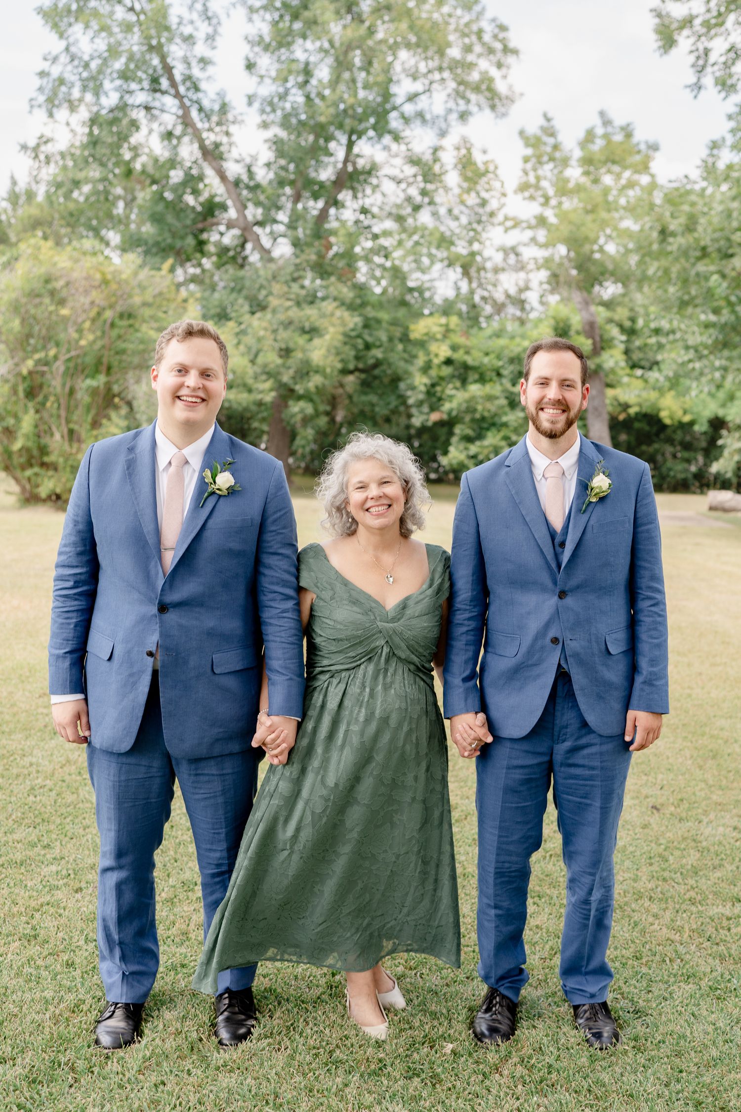 Wedding party members in navy suits and a green dress pose together on grass under trees.