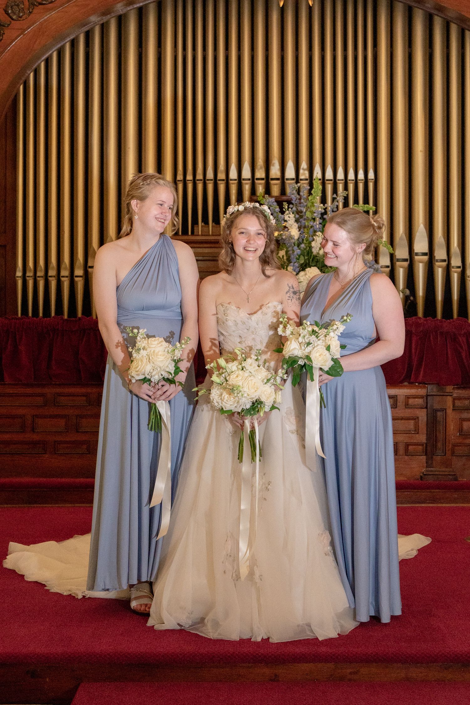 Three people in formal attire stand on red carpeted church steps with organ pipes in background, holding floral bouquets.