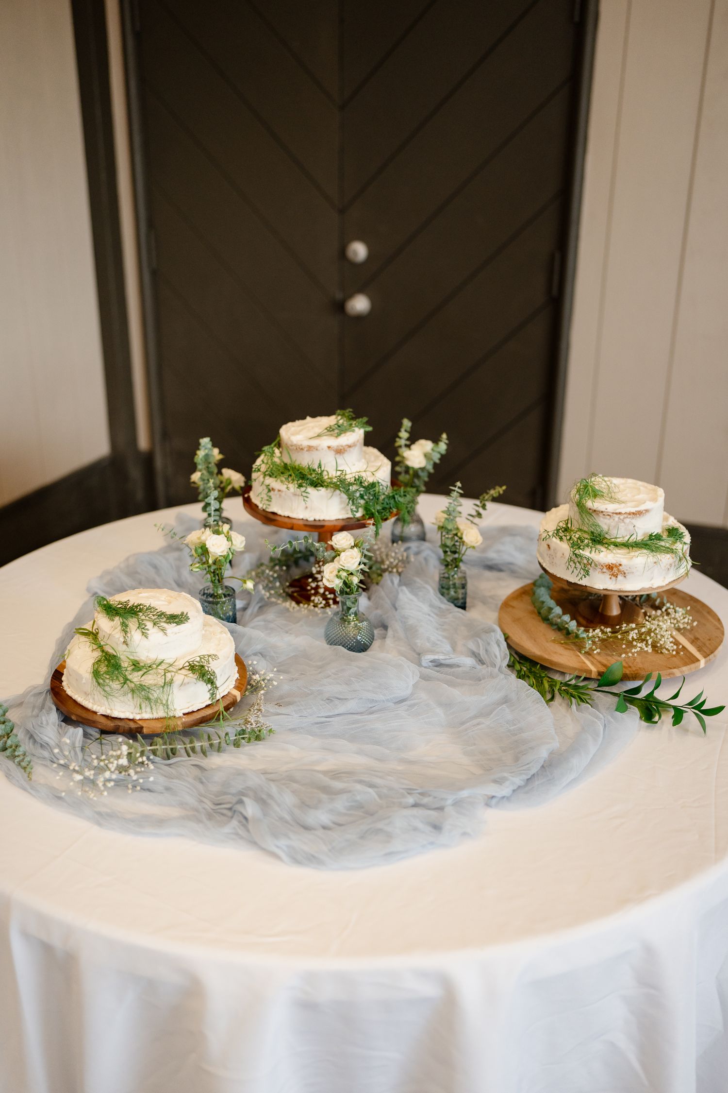 Three decorated wedding cakes displayed on white tablecloths with delicate floral and greenery arrangements.