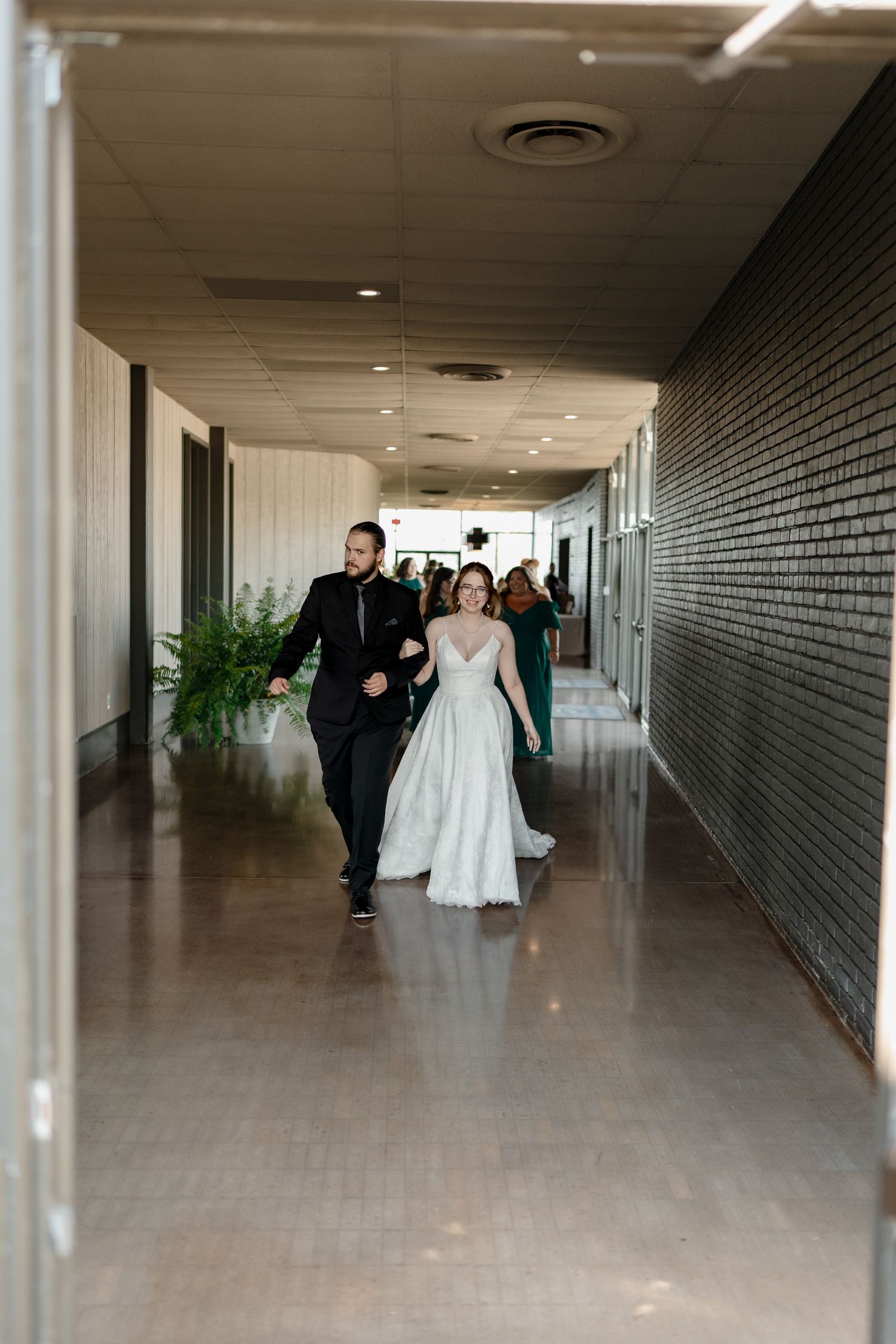 Wedding party walks through a long hallway with grey brick walls and white ceiling lights.
