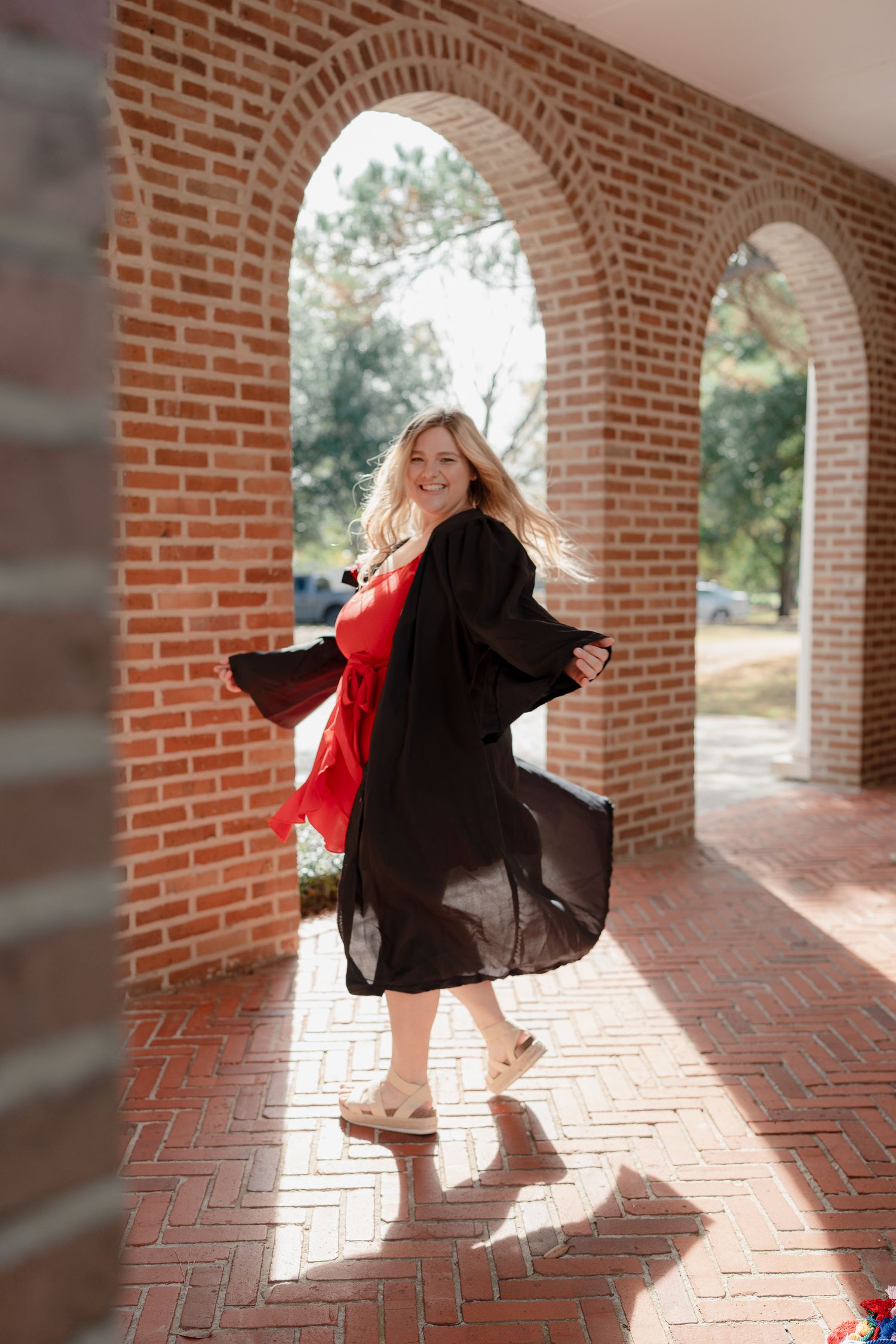 Dancing figure in a black graduation gown and red dress twirls beneath brick archways on a sunny day.