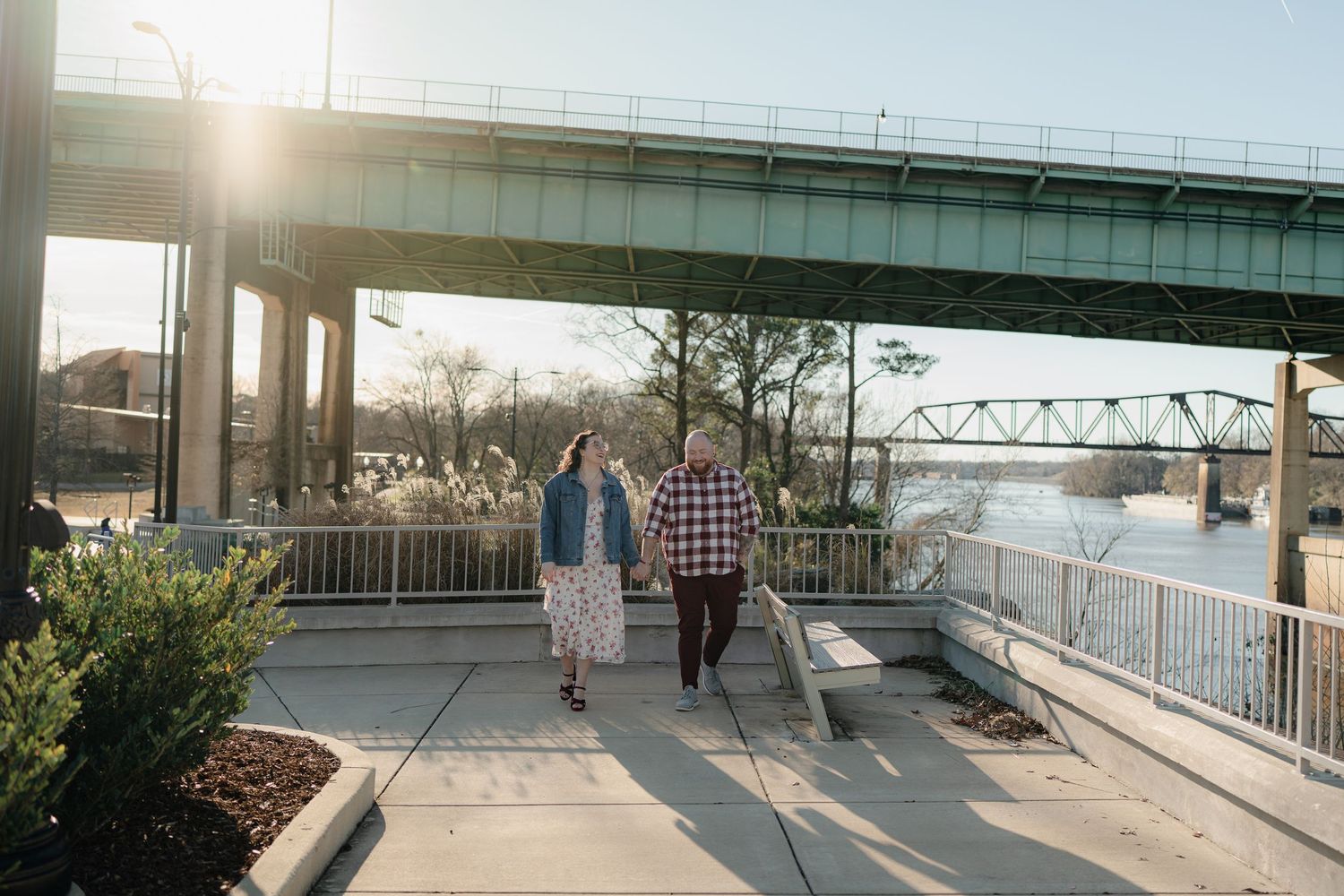 Couple walking along a riverside path with bridges and buildings in the background during sunset.
