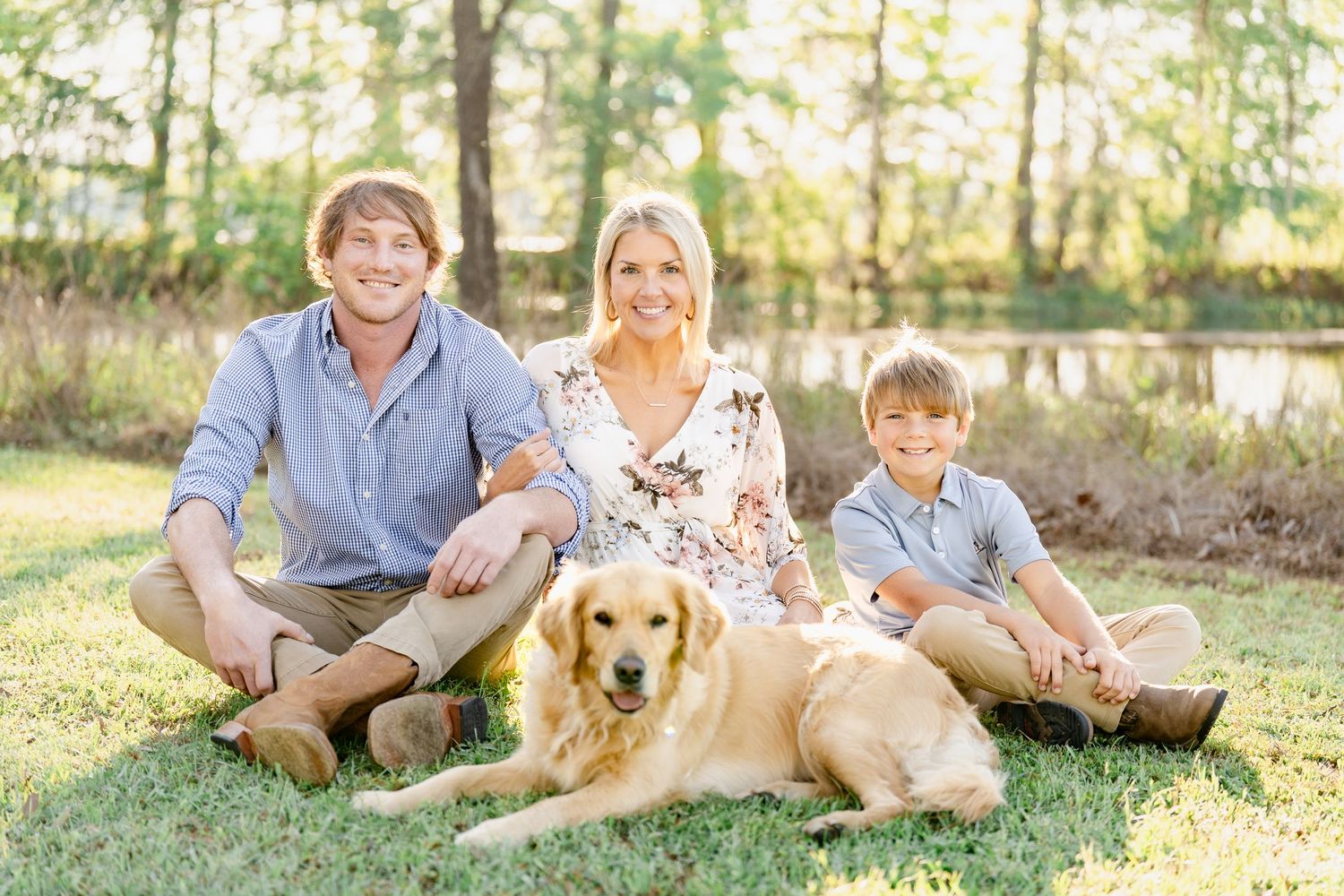 Family and dog relax together on grass in sunny wooded setting during outdoor photo session.