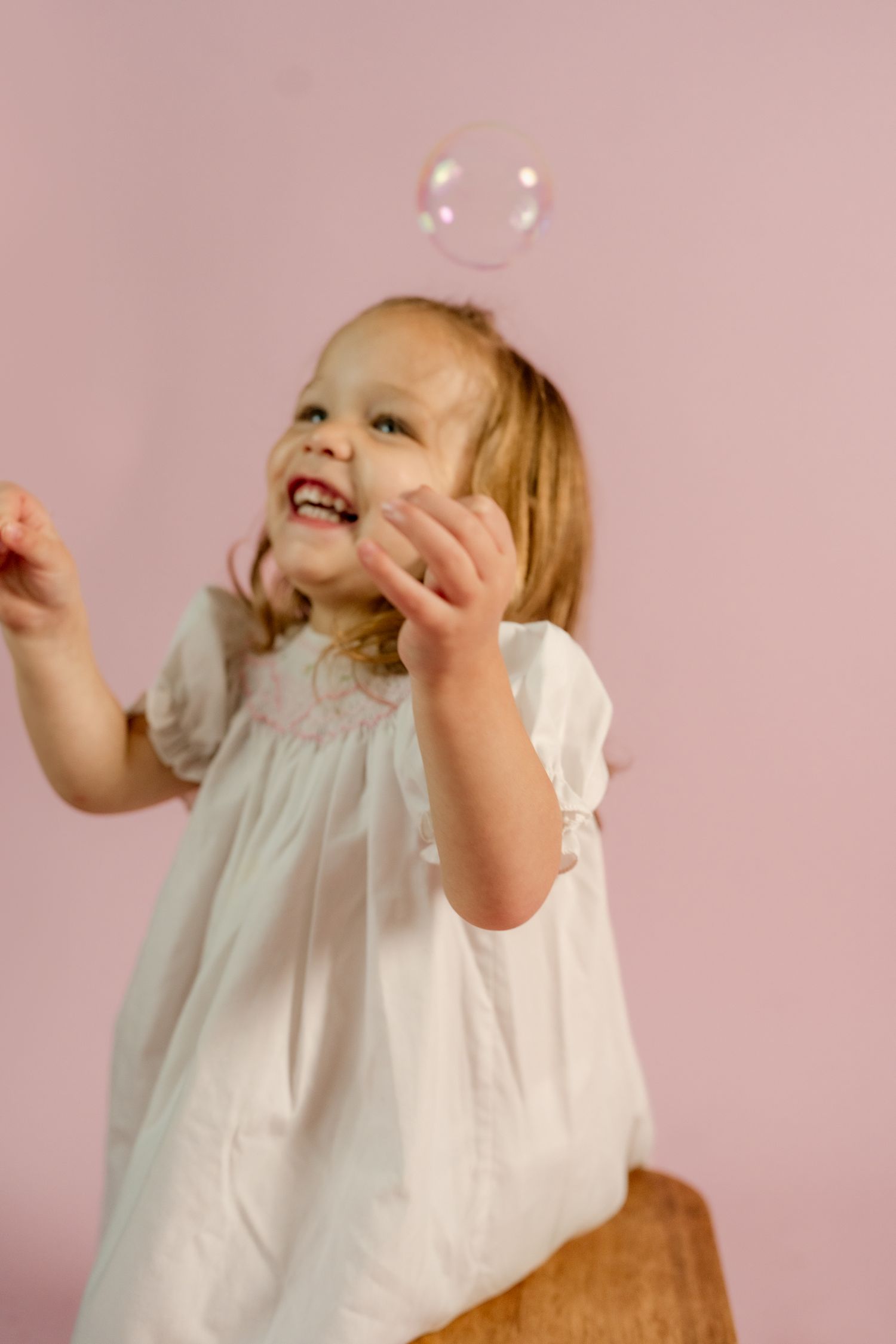 A joyful moment captured as a child in white plays with bubbles against a pink backdrop.