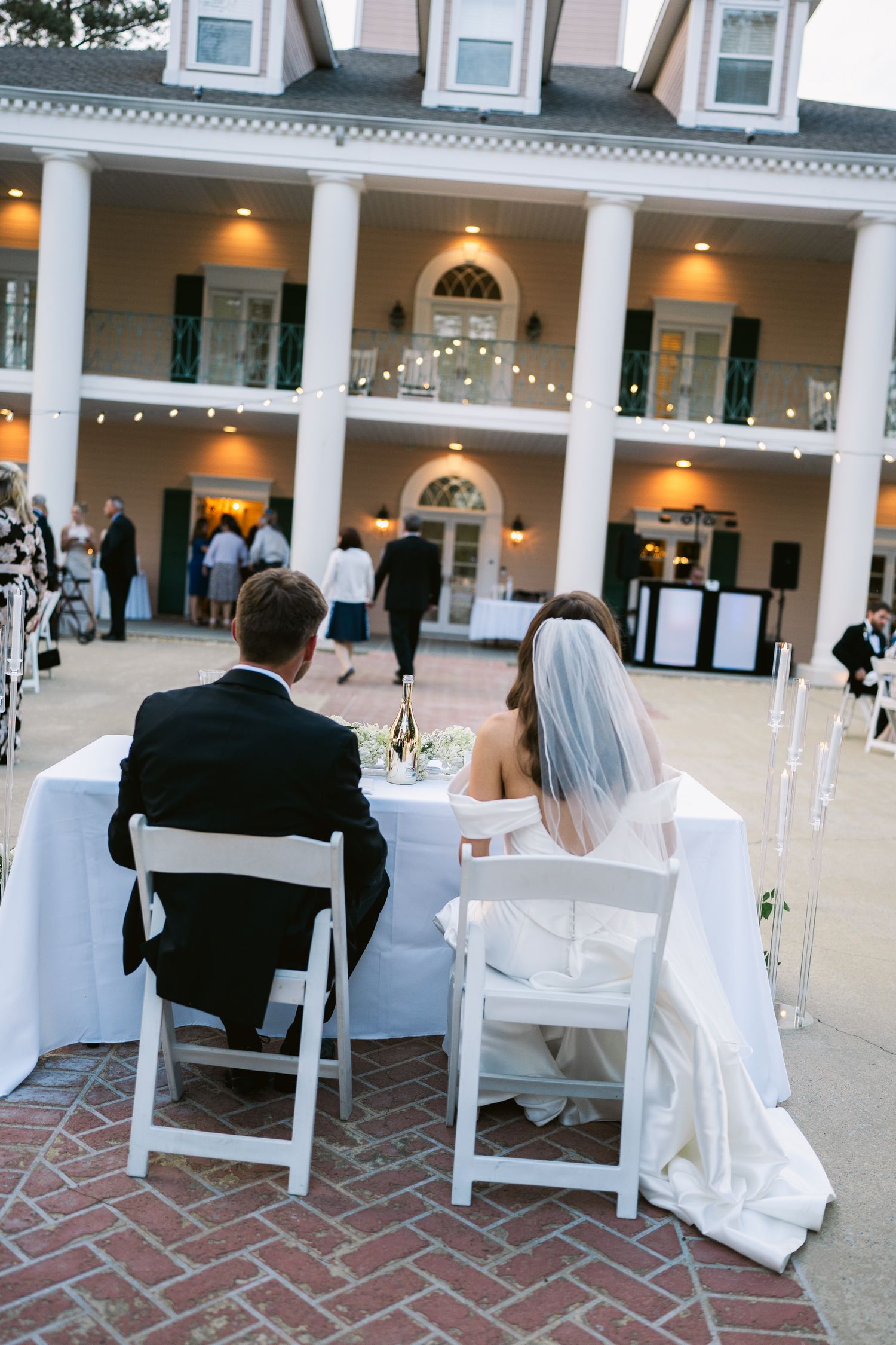 Wedding couple seated at reception table in front of illuminated Southern mansion.