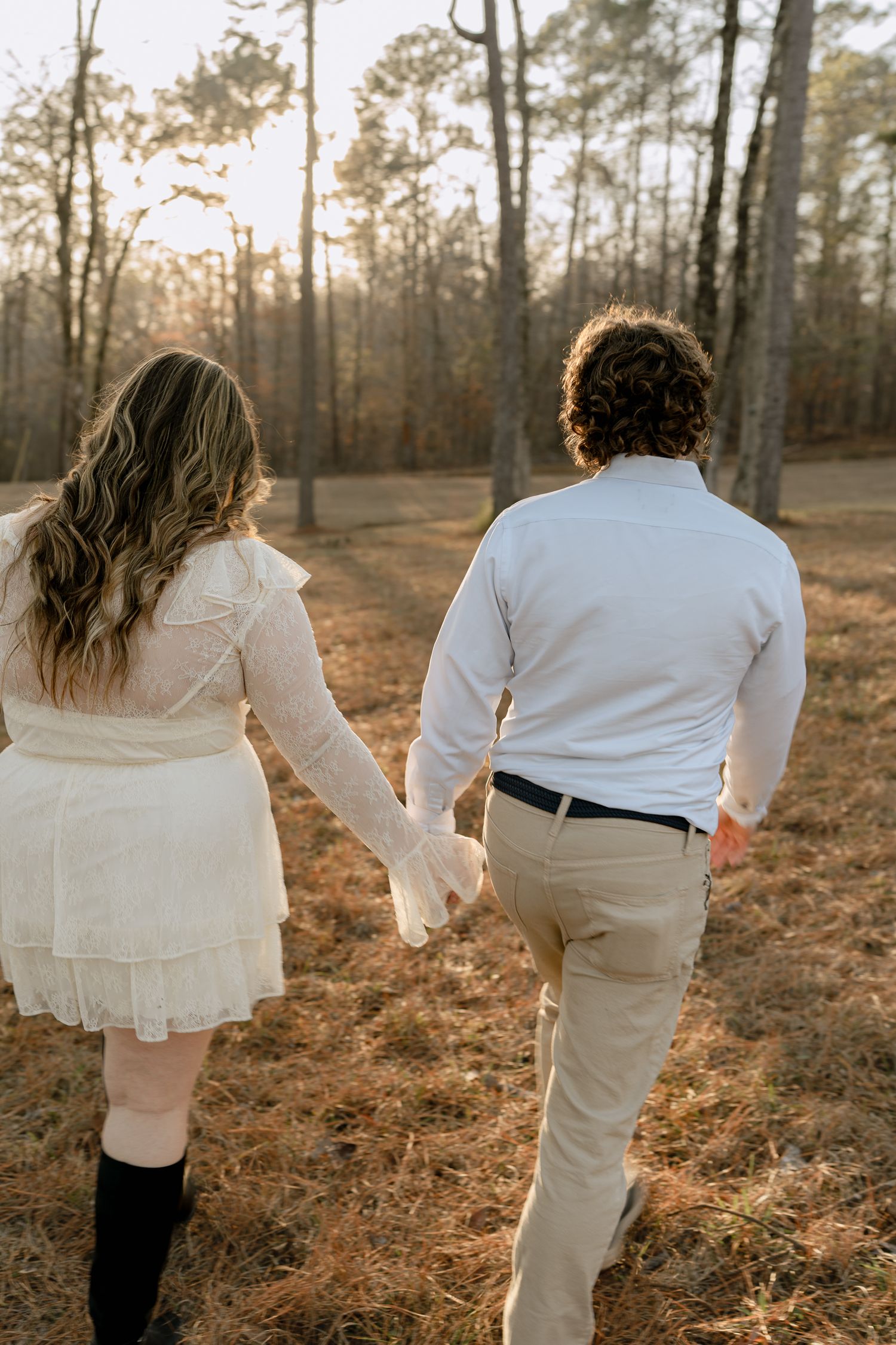 Couple walking hand in hand through a pine forest at golden hour, viewed from behind.