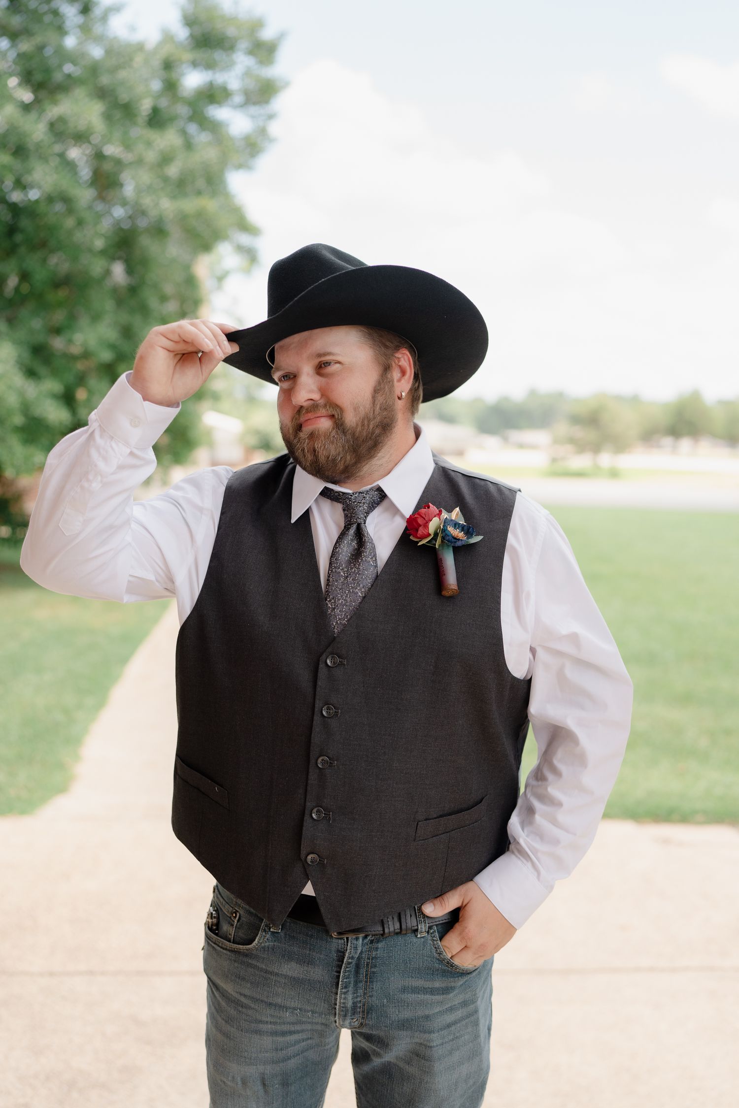 A western-styled groomsman in a dark vest, white shirt and cowboy hat poses outdoors on a dirt path.