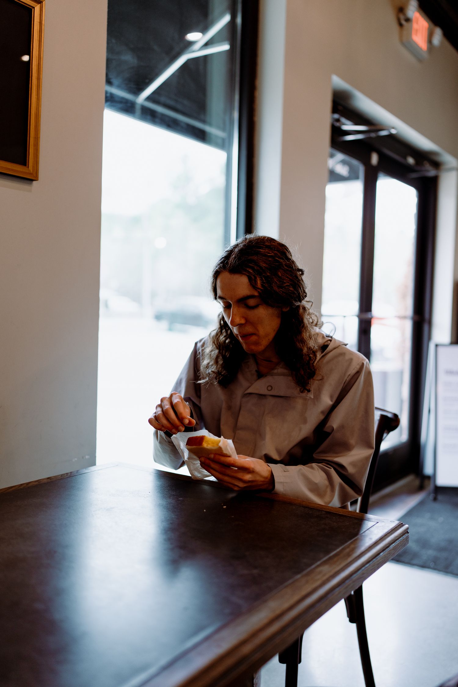 Individual in light colored jacket looking down at food while seated at dark table.