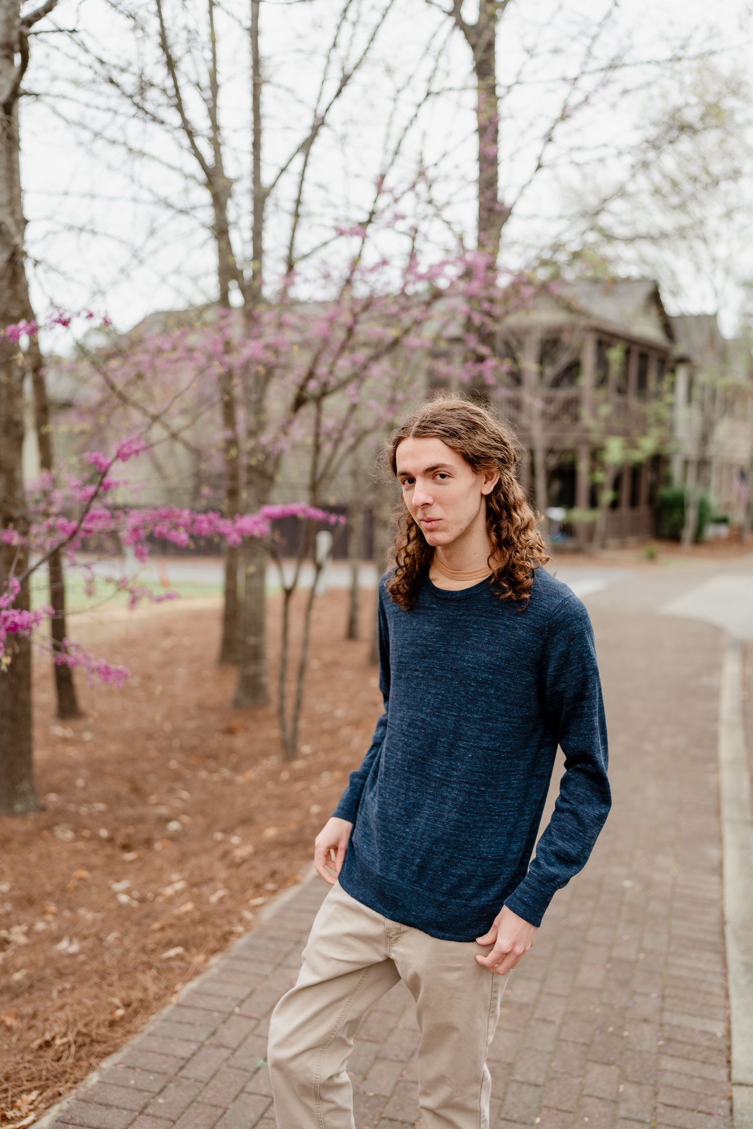 Person in navy sweater standing on brick pathway with flowering trees and buildings in background.