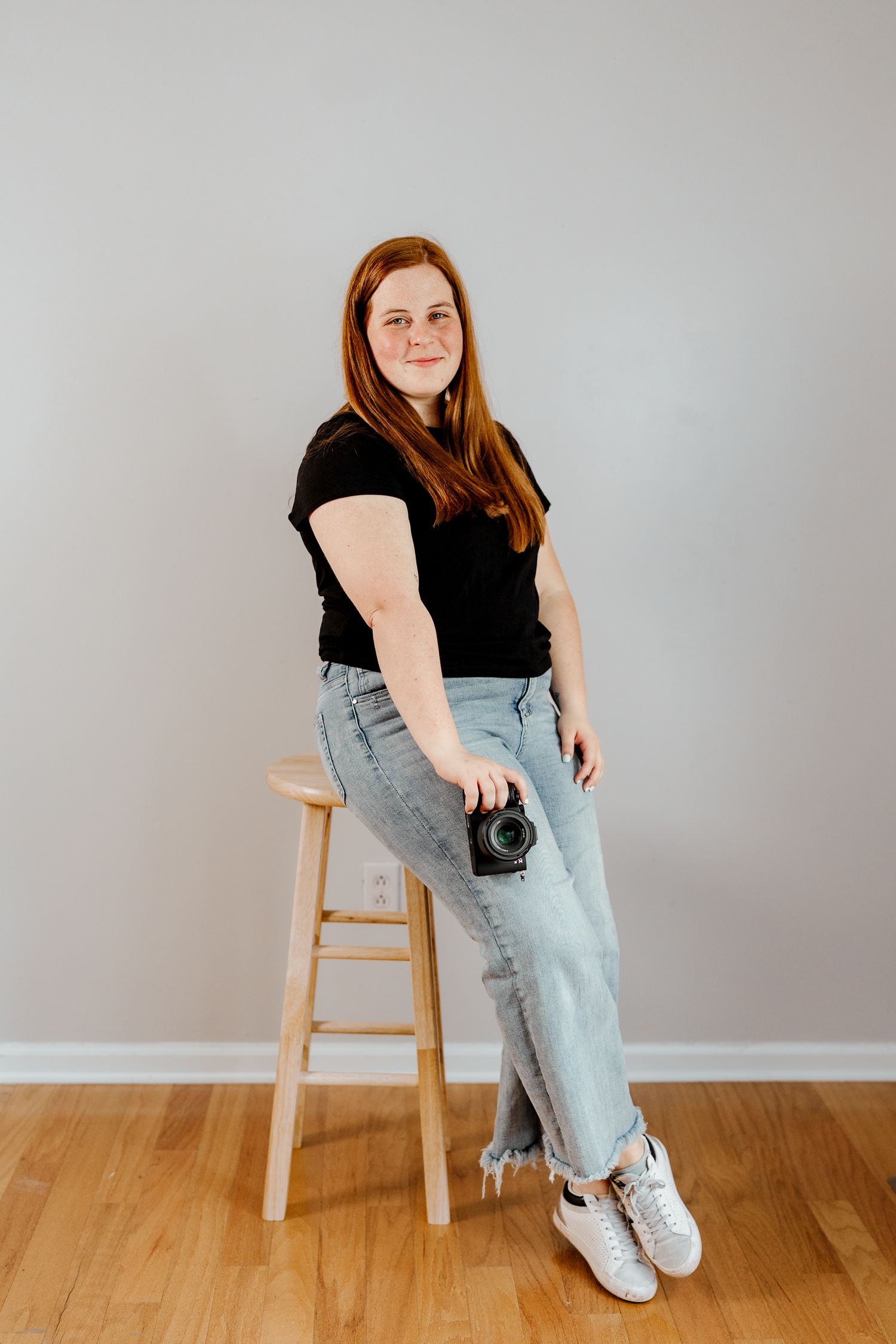 Person in black top and light jeans sits casually on wooden stool against grey wall with white sneakers.