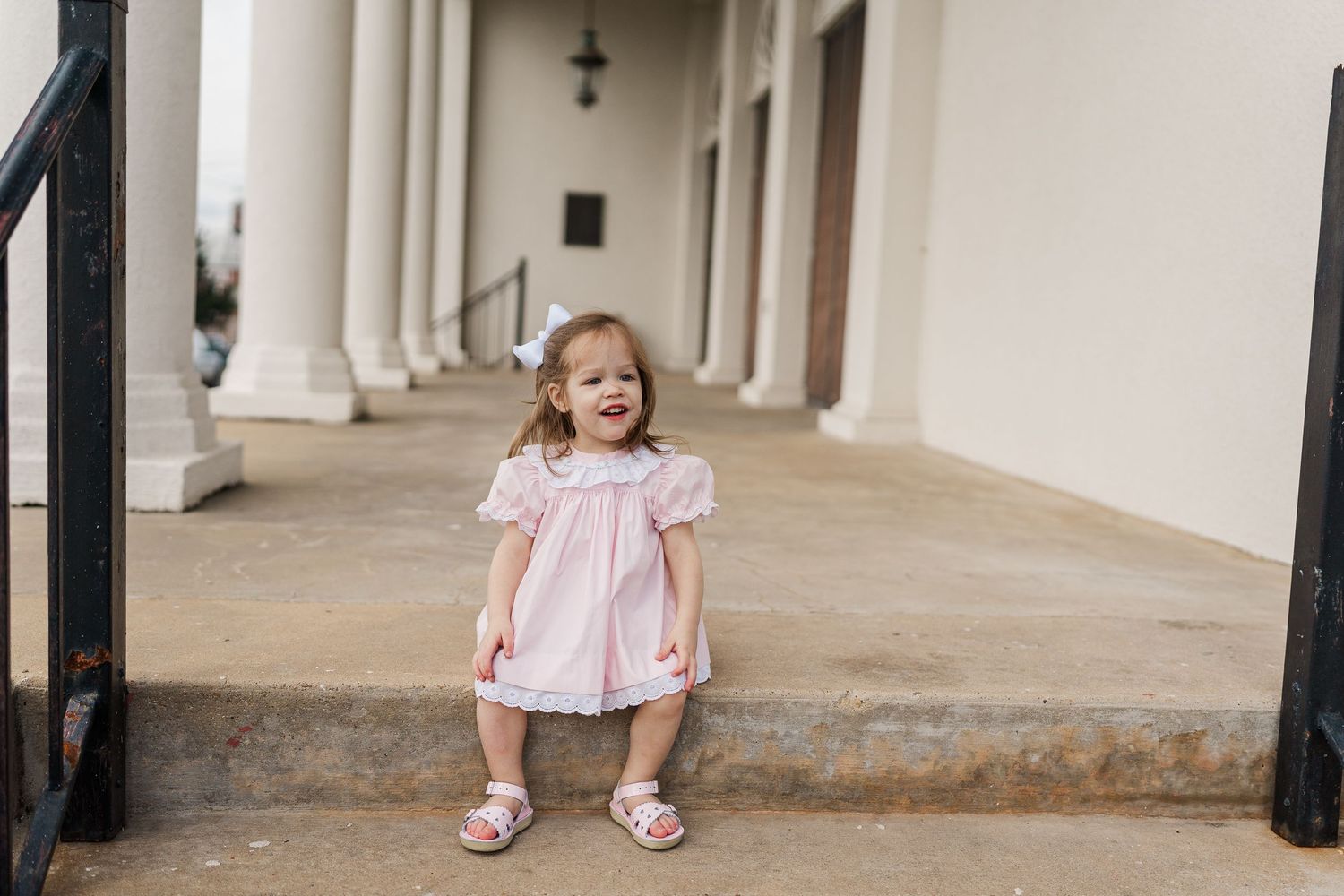 A little one stands between white columns wearing a pink dress and sandals.