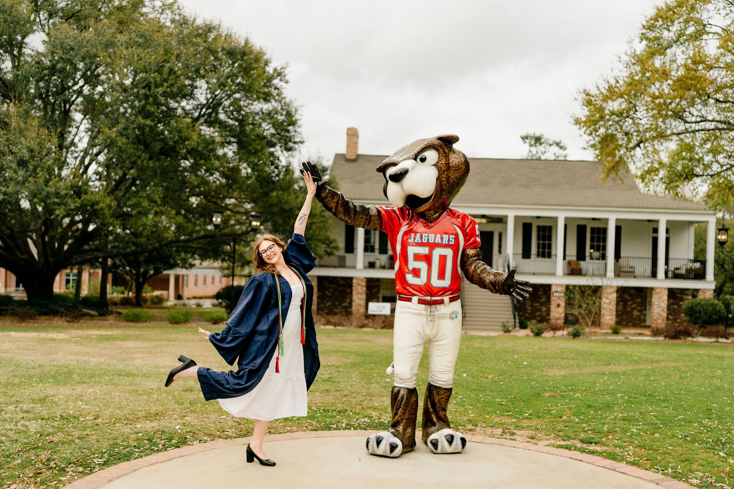 Graduate in white dress and academic regalia poses with university mascot on brick pathway.