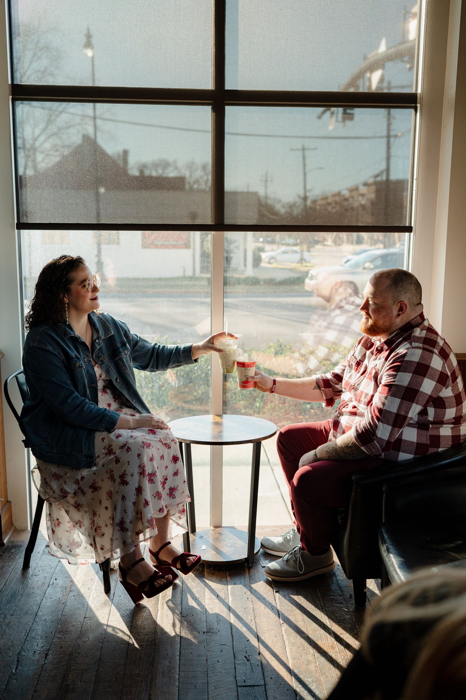 Two people sit by large windows in a cafe sharing conversation and coffee in natural sunlight.