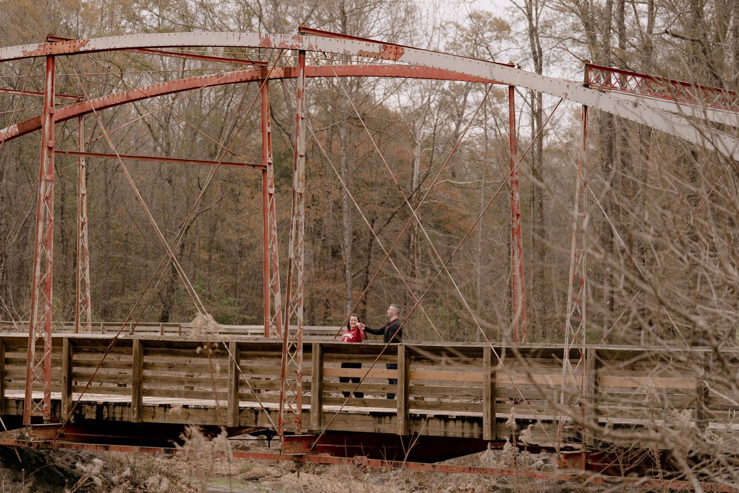 A distant view of a couple on a red-painted metal bridge surrounded by winter trees.