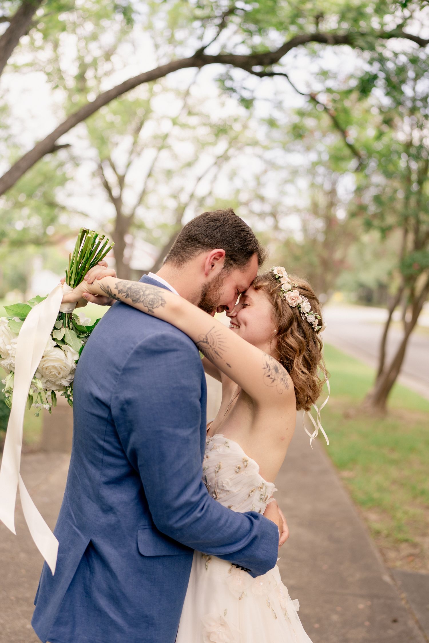 Newlyweds embrace and share intimate kisses under green trees on a garden pathway.