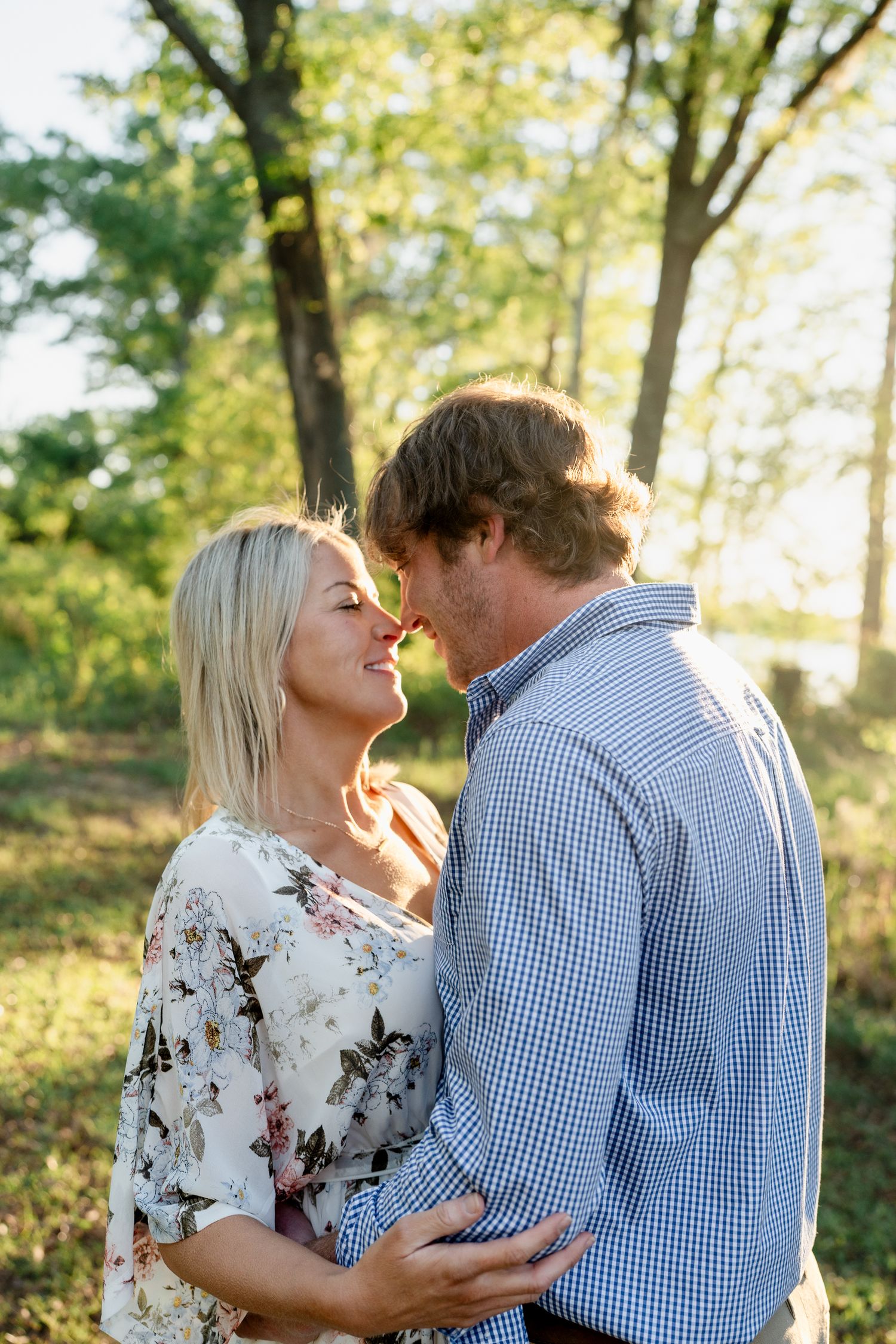 A couple shares an intimate moment in dappled sunlight with trees in the background.
