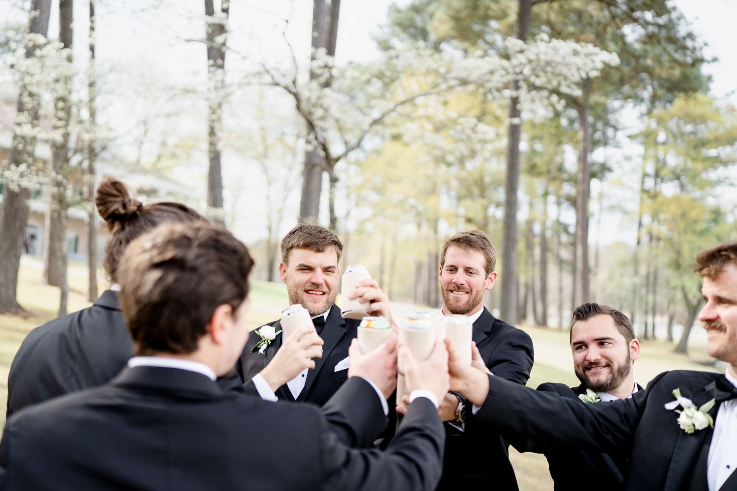 Groomsmen in black tuxedos toast and celebrate outdoors among pine trees.