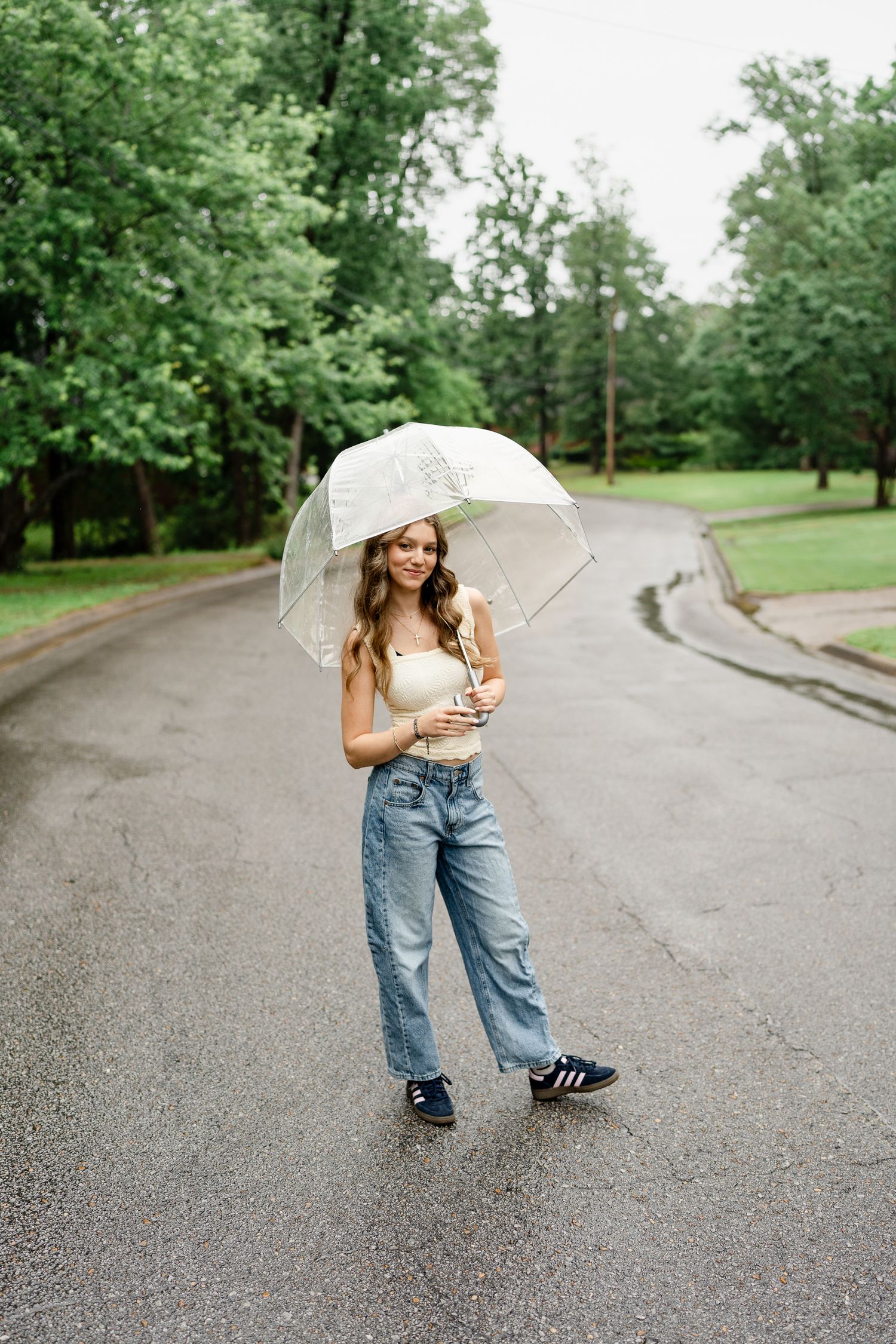 Full length shot of casual outfit with clear umbrella on wet road surrounded by green trees.