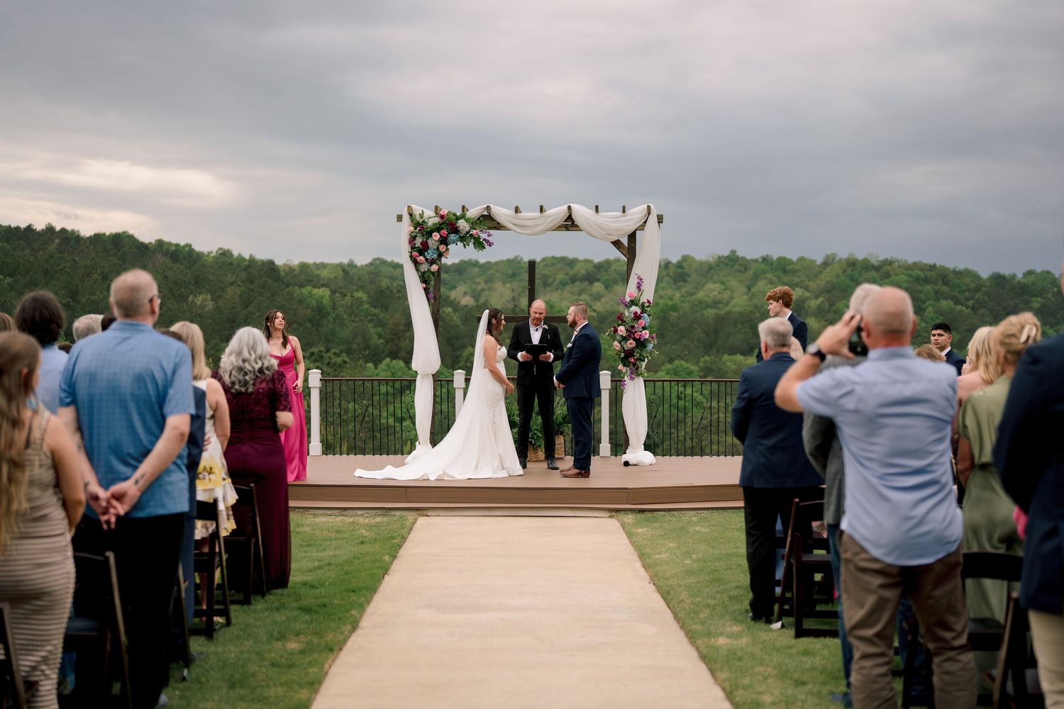 Outdoor wedding ceremony with bride and groom at floral arch altar, guests seated on green hillside.