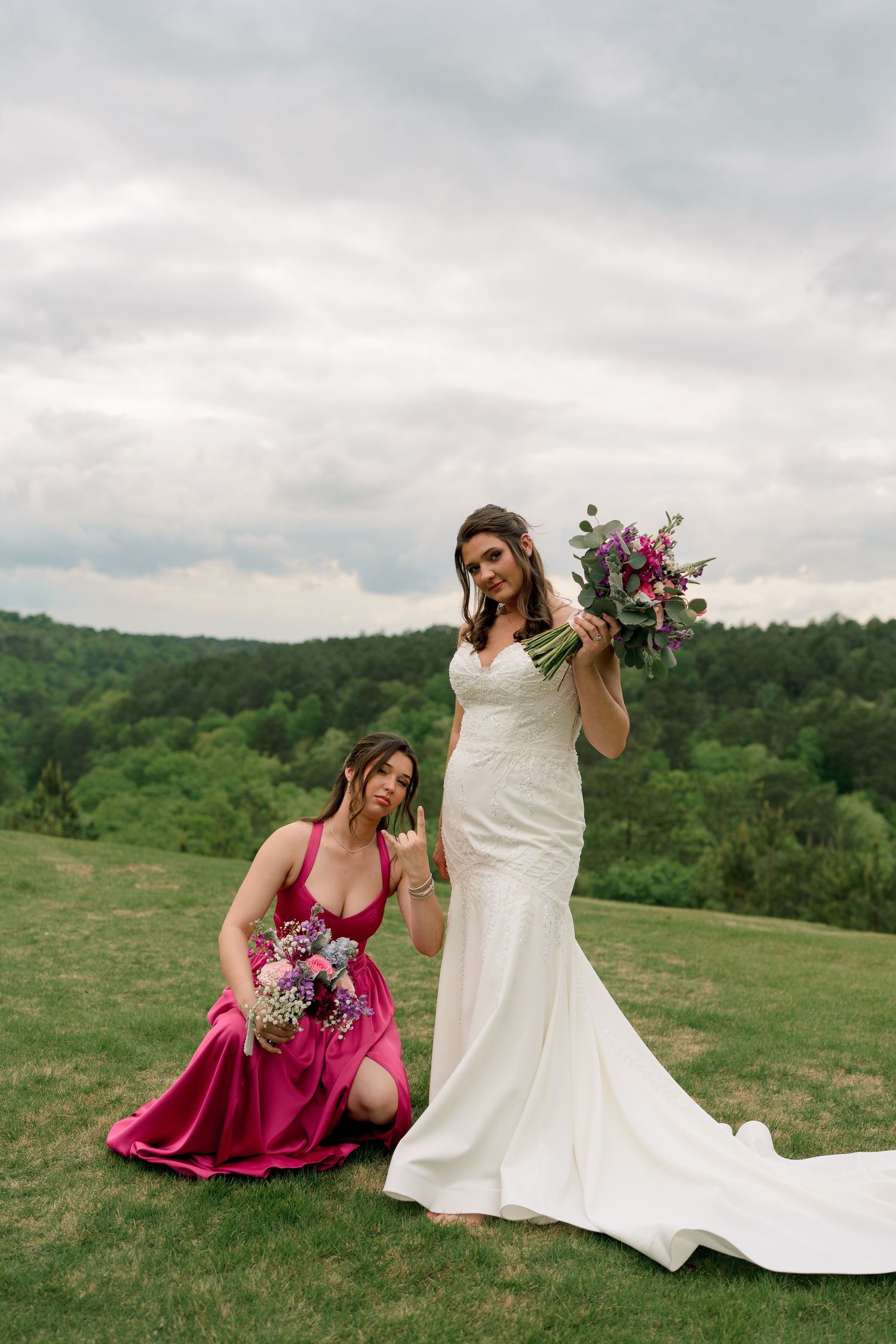 Bride in white gown poses with bridesmaid in pink satin dress holding purple floral bouquets outdoors.
