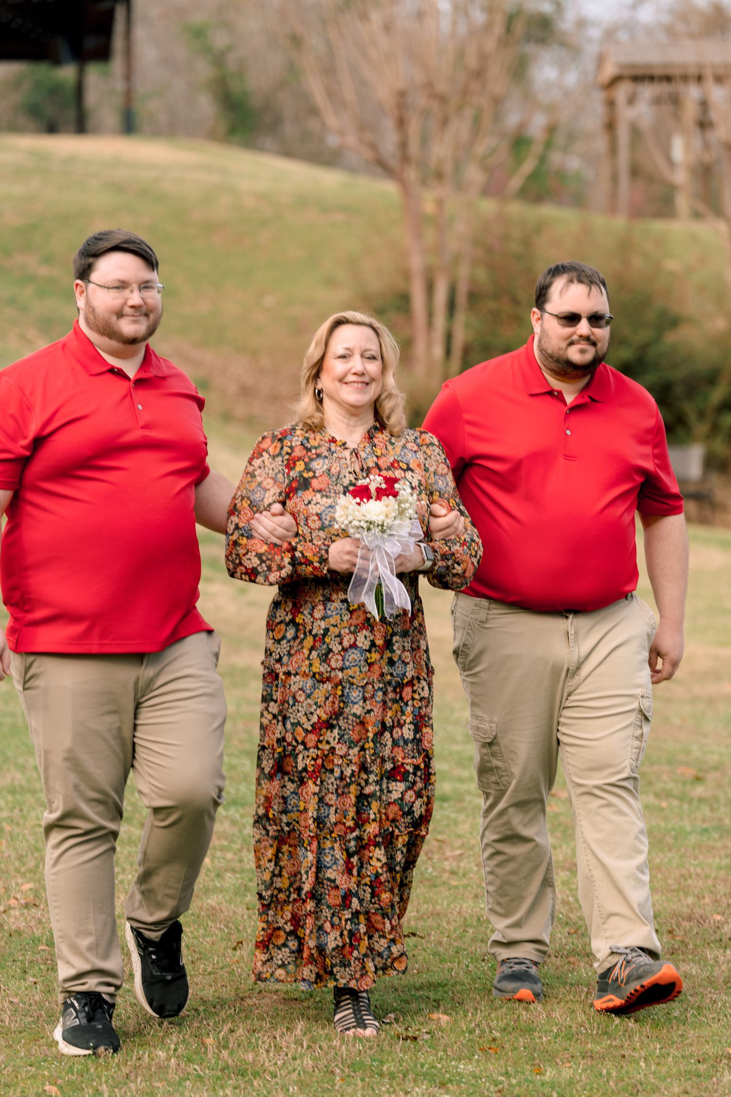 Two men in red polos escort a smiling woman in a floral dress holding a bouquet outdoors.