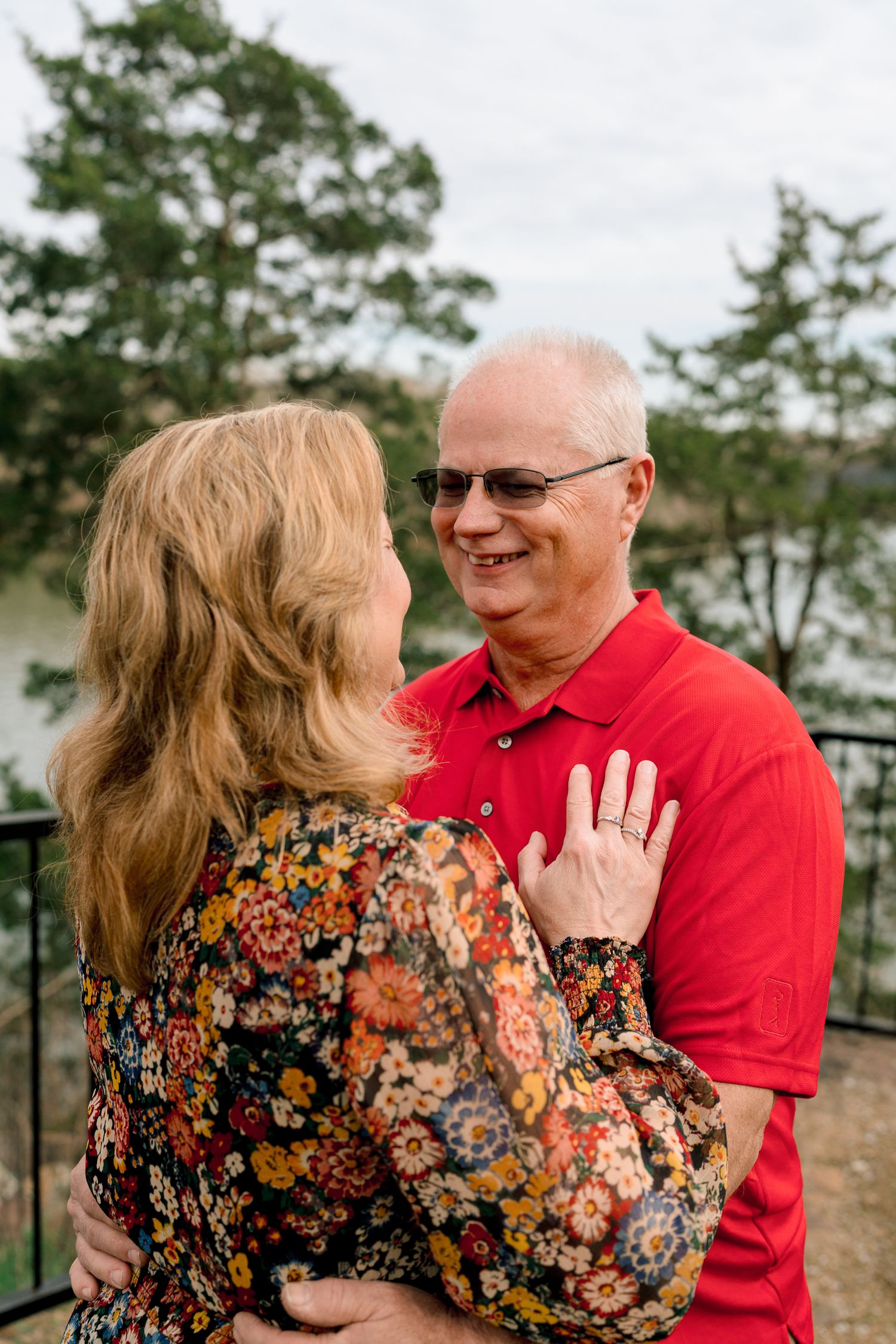 Smiling older man in red polo embracing a woman in a floral dress with a lake view behind them.