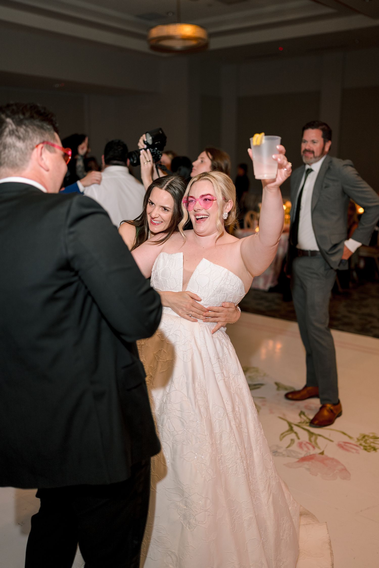 Bride in white gown raises glass while dancing with guests at elegant wedding reception celebration.