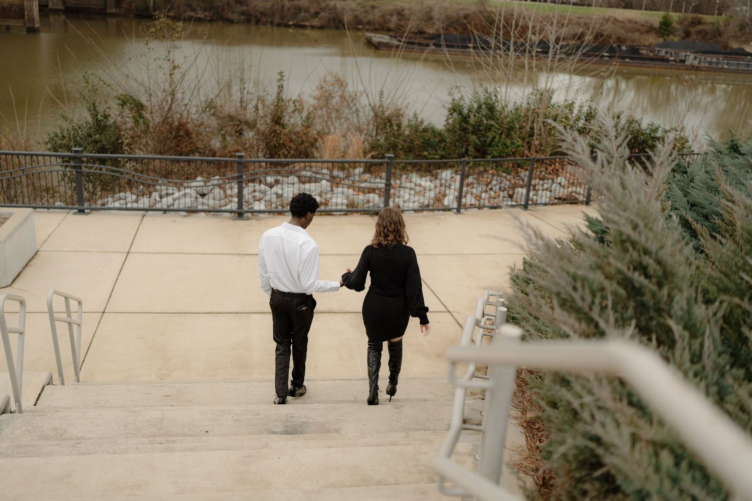 A couple walks hand in hand along a riverside concrete path surrounded by autumn foliage.