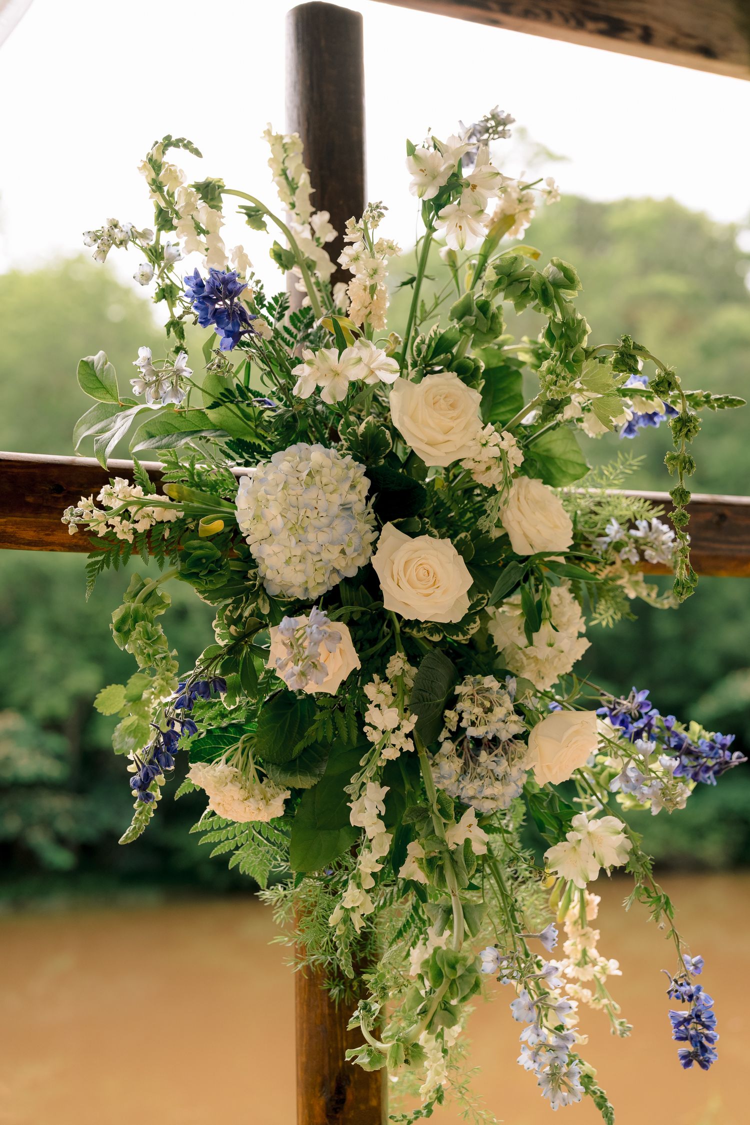 Elegant wedding arch floral arrangement with white roses, hydrangeas, and blue wildflowers on rustic wood.