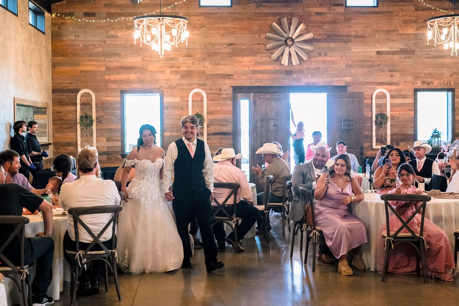 Bride and groom making grand entrance into rustic barn wedding reception with chandelier lighting.