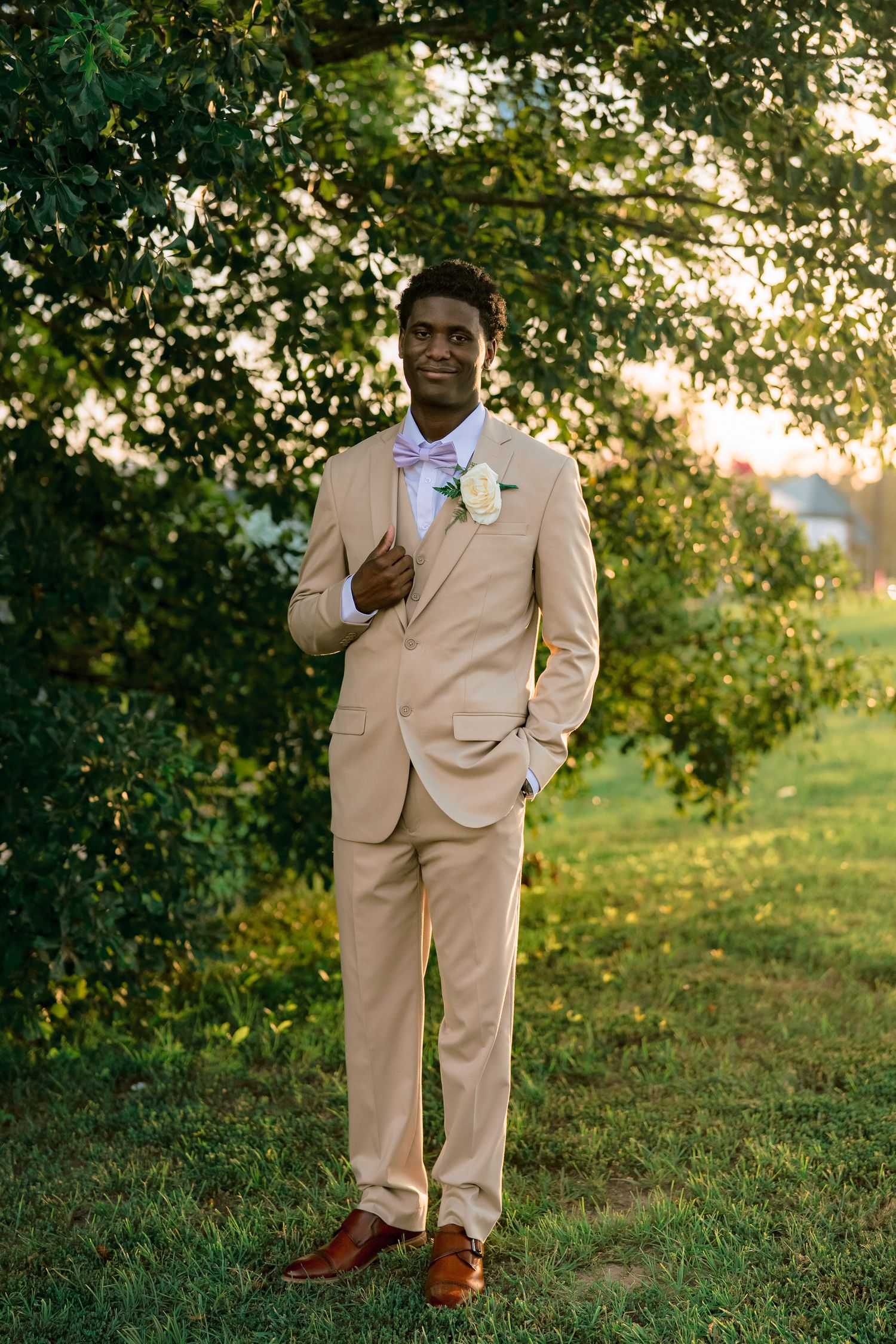 Handsome groom in tan suit with boutonniere poses under green tree in golden sunset light.