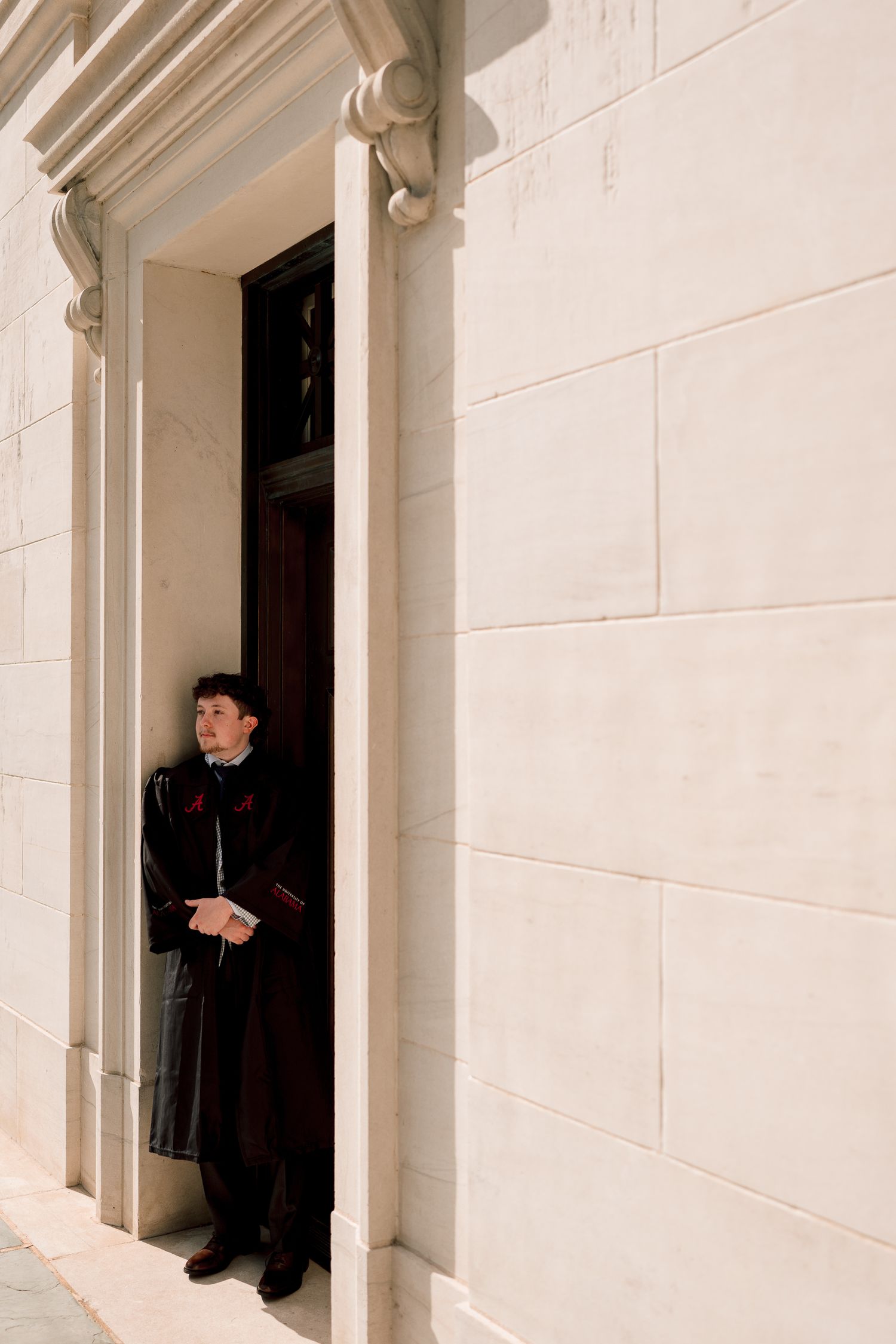 Young man in dark formal attire standing in doorway of classical stone building holding a book.