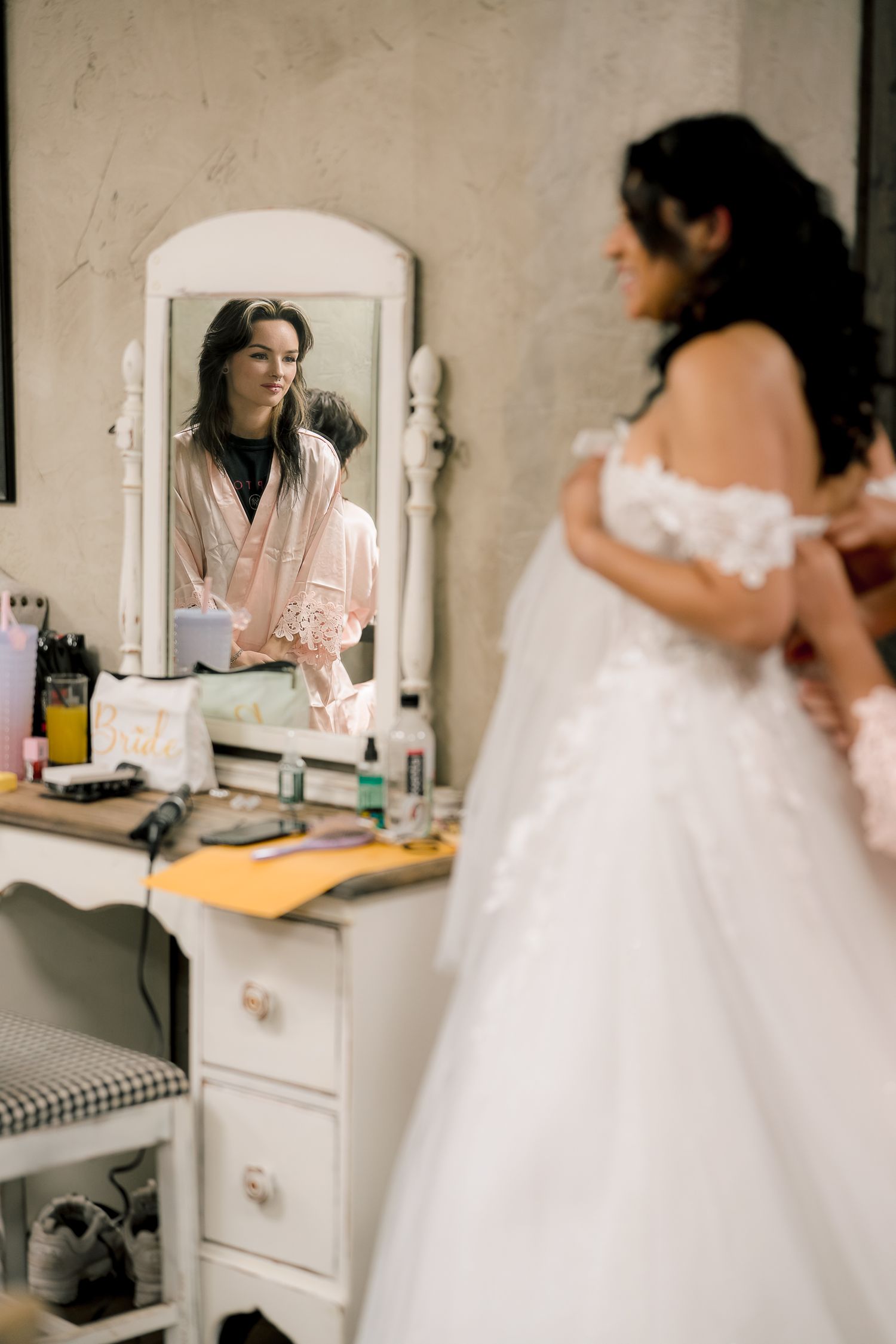 Bride in white lace wedding gown getting ready, reflected in vintage vanity mirror with bridesmaid in pink robe.