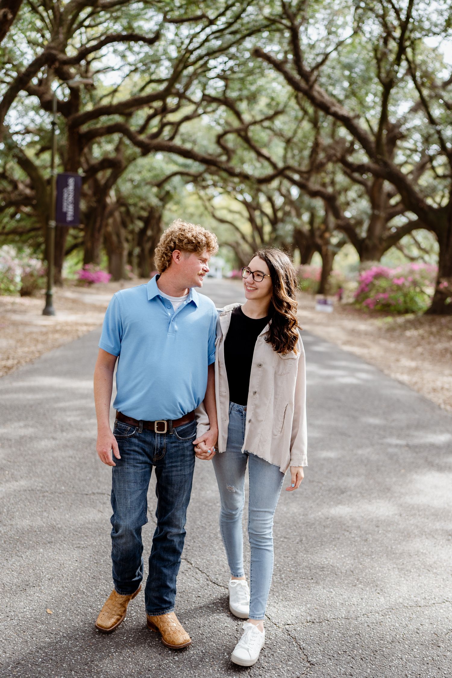 A couple walks along a path under oak trees on a sunny day.